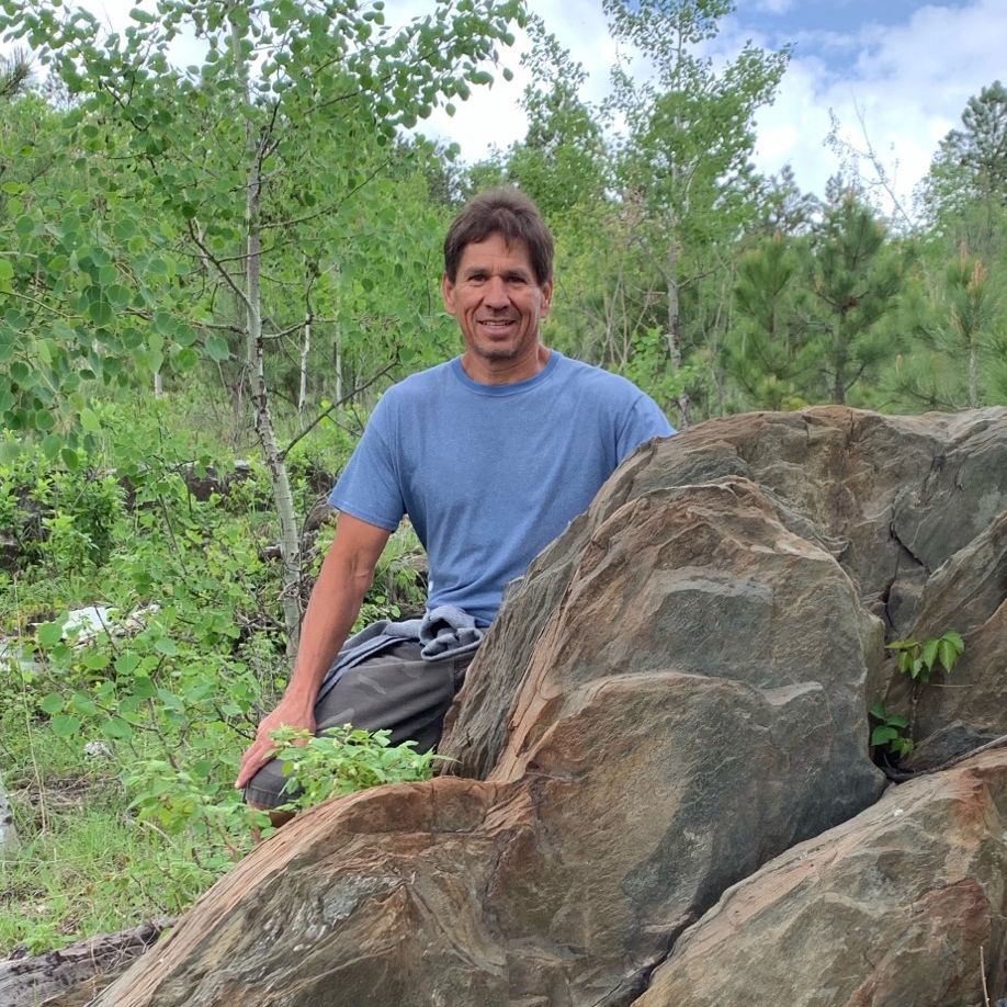 Man kneeling next to a large rock formation, wearing blue shirt, outdoor setting with trees.