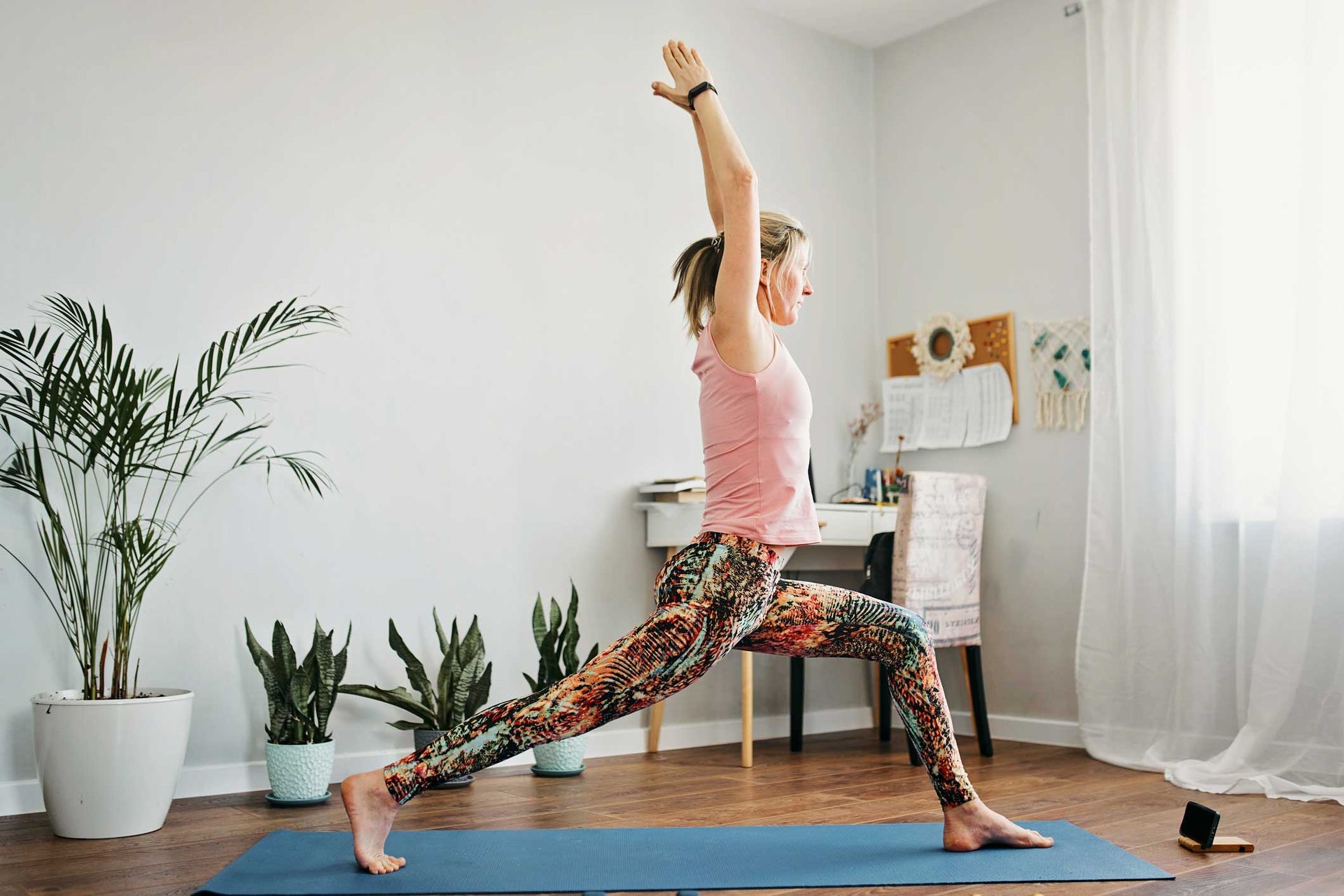 Woman in yoga pose, arms raised, in a brightly lit room with plants and a desk.
