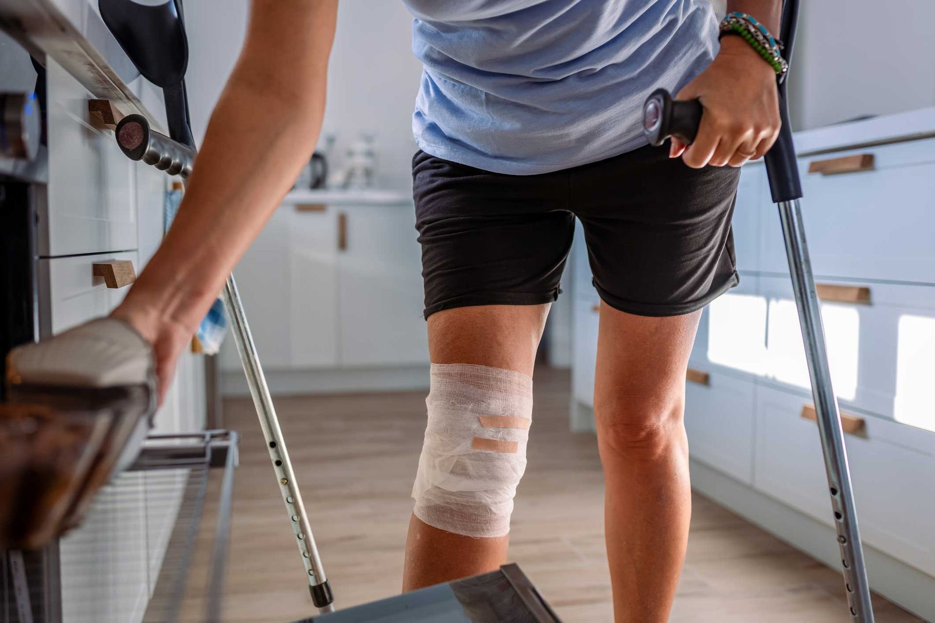 Person with bandaged knee uses crutches and holds onto furniture for support in a kitchen.
