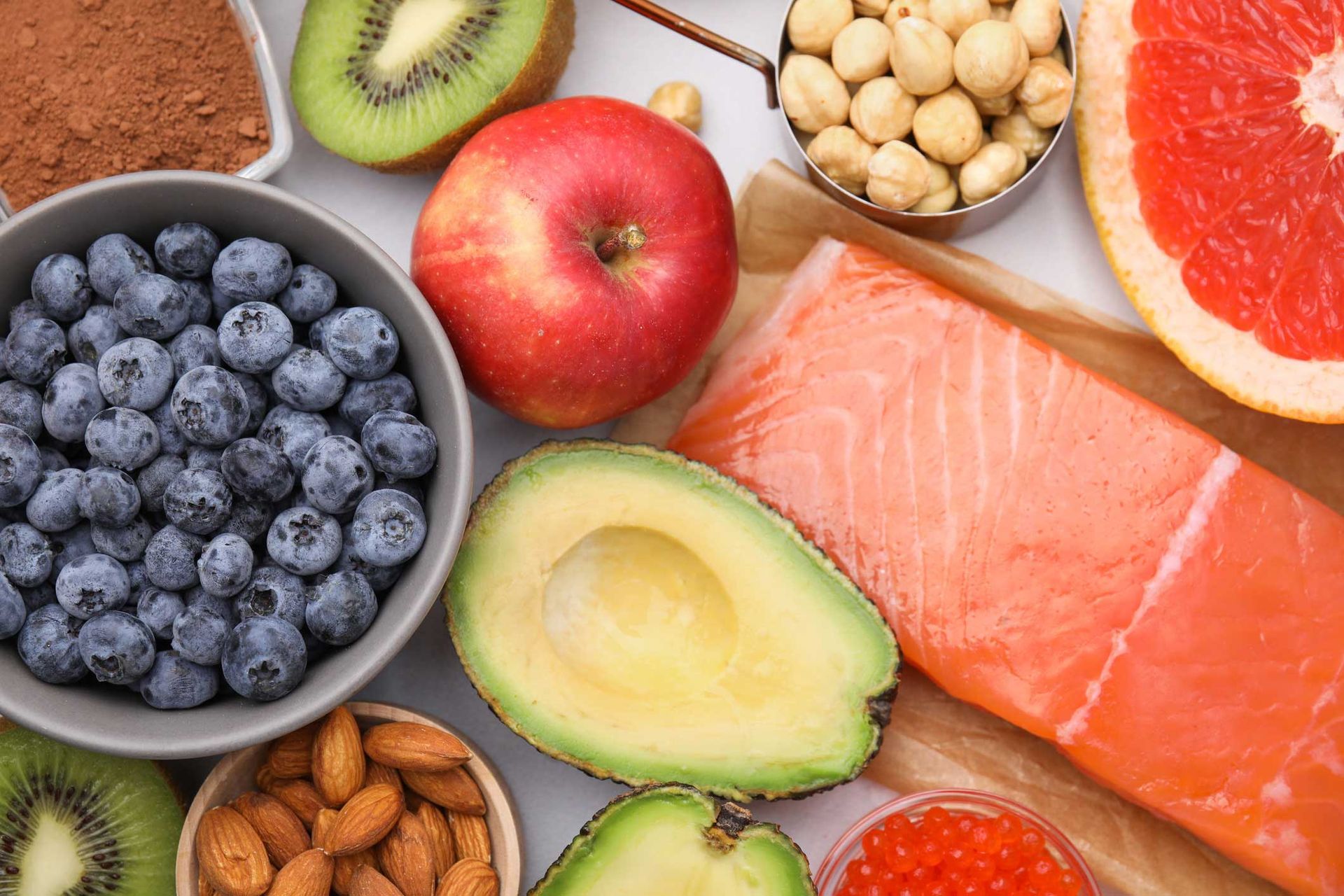 Assortment of colorful healthy foods: blueberries, apple, salmon, avocado, kiwi, nuts, grapefruit.