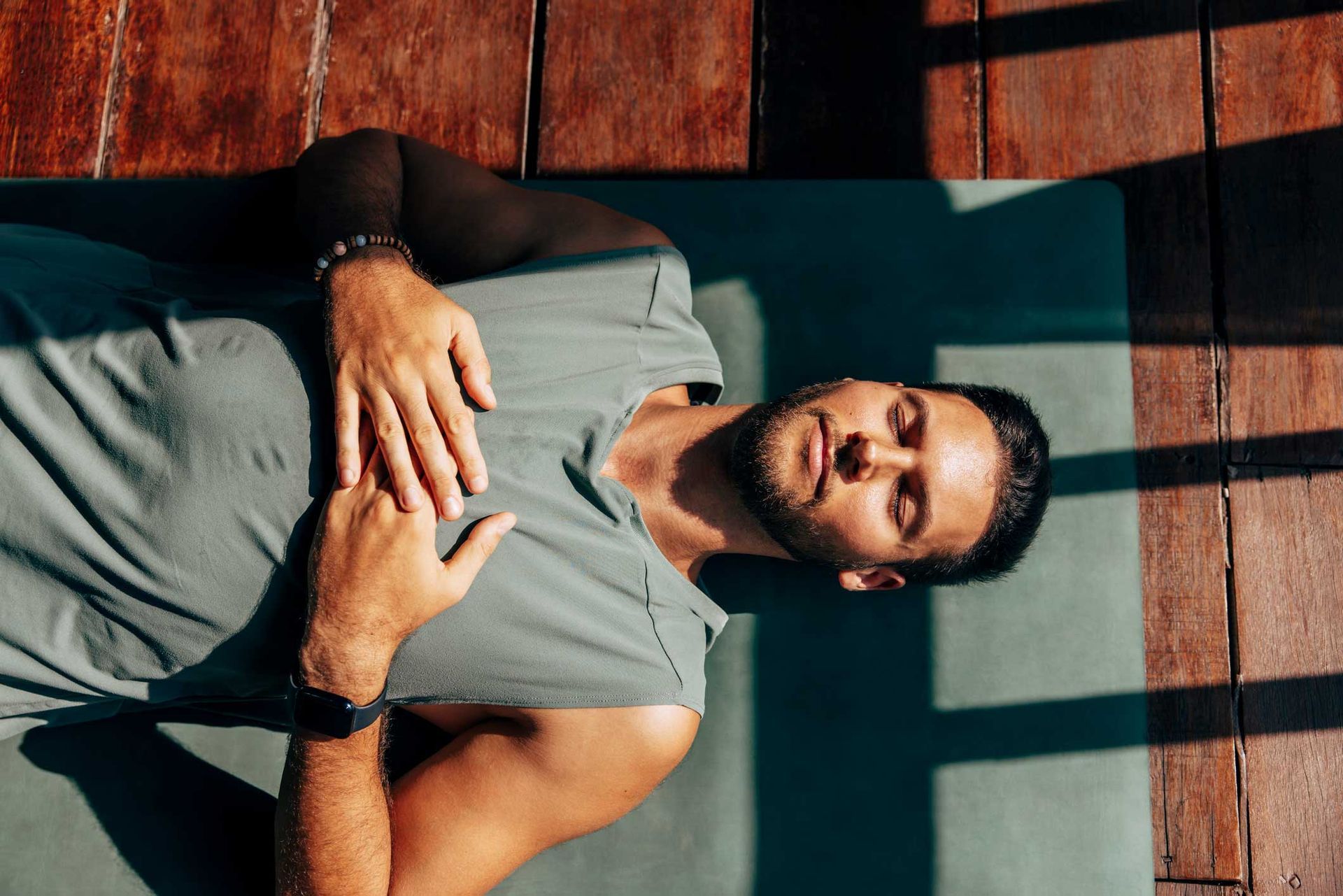 Man lying on a mat outdoors, hands on chest, eyes closed, sunlit.
