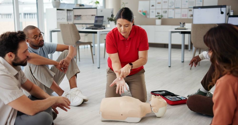 CPR instructor demonstrating on a dummy, surrounded by seated students in a classroom.