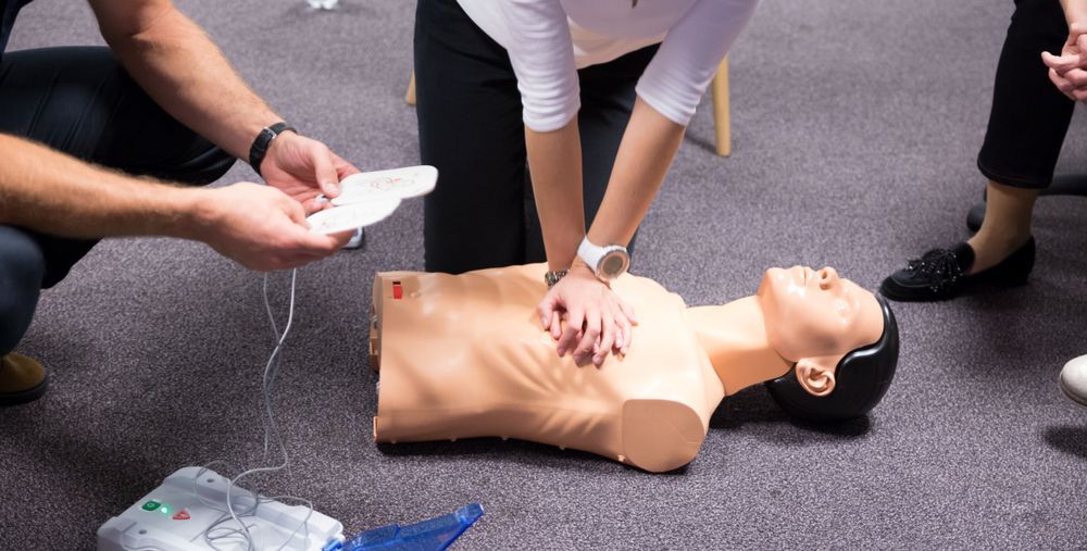 People practicing CPR on a mannequin, with one person holding an AED pad in a classroom setting.