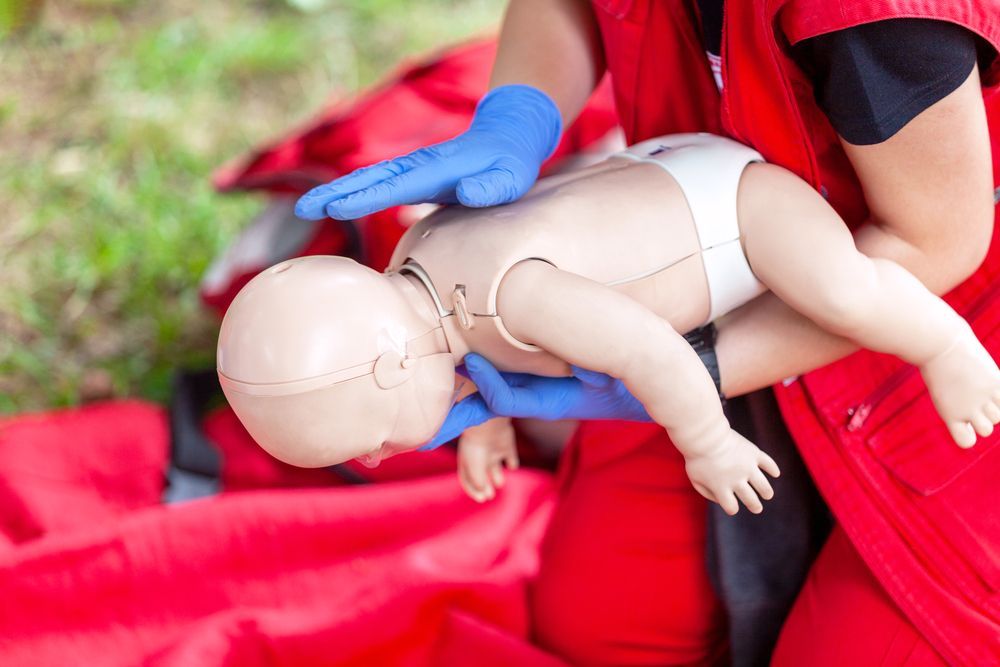 Person in red vest demonstrates infant CPR on a baby mannequin, outdoors.