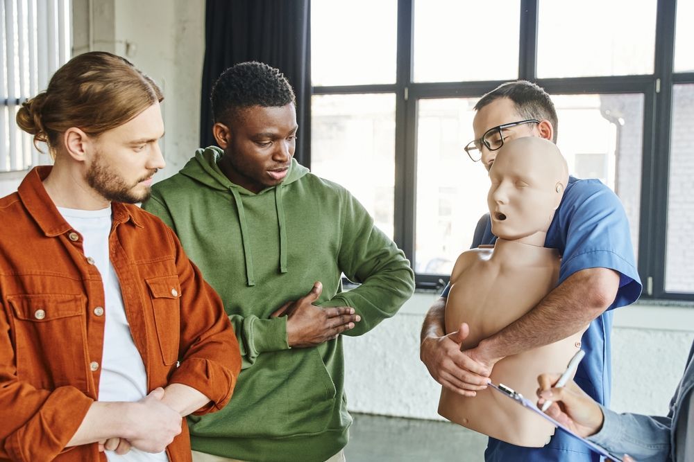 Four people in a room during a CPR training session; instructor holding a mannequin, others observing.