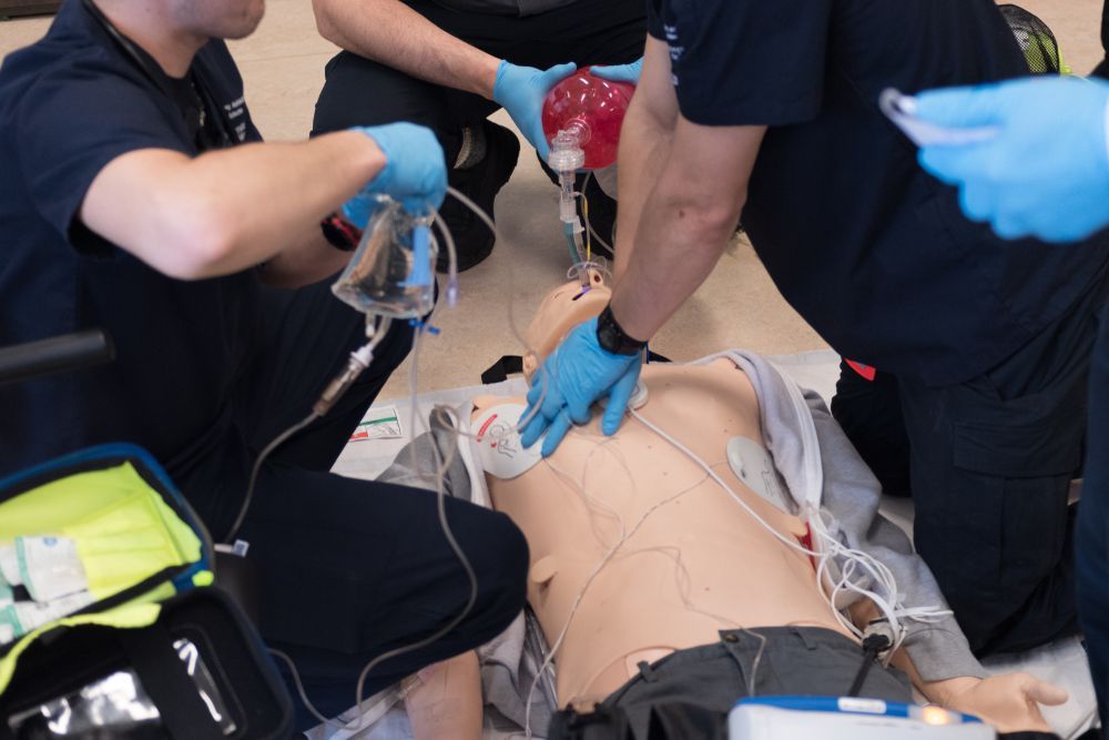 Medical personnel practice CPR on a mannequin, using an Ambu bag, with equipment in a training setting.