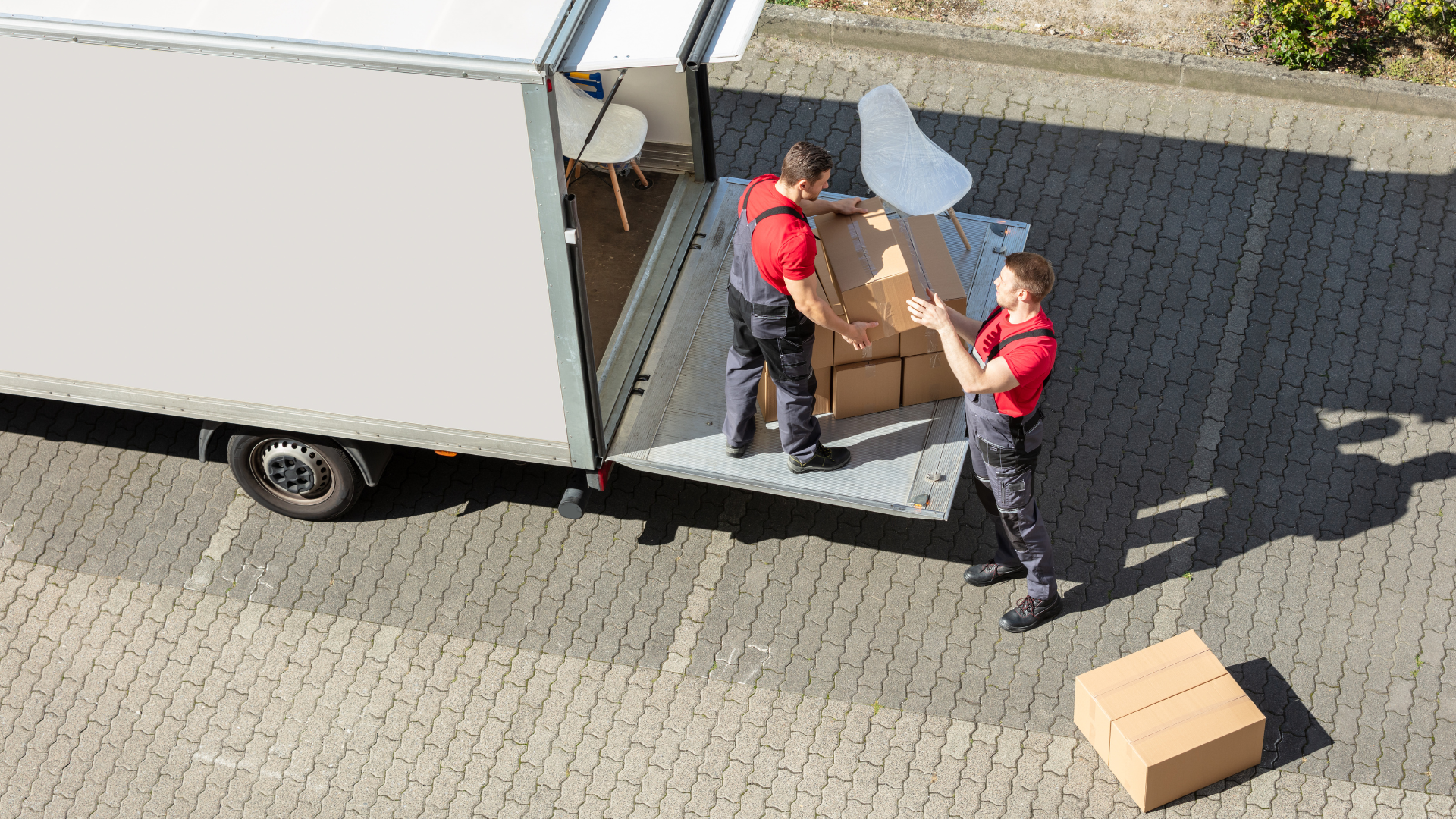 Two movers loading a box into a truck, one catching and the other directing, on a brick parking lot.