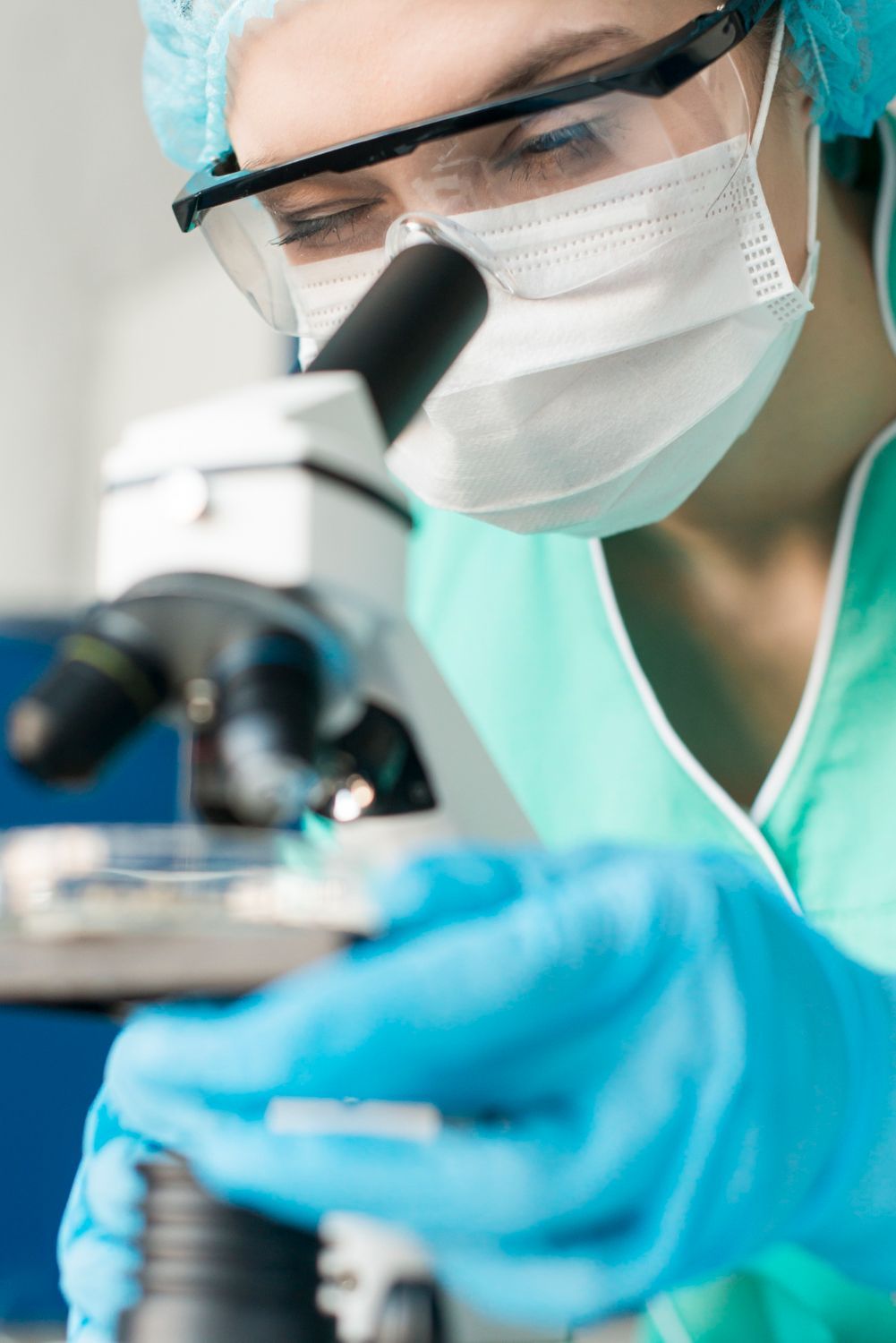 Un científico con gorro médico, mascarilla y guantes azules examina una muestra en una lámina de microscopio en un laboratorio.