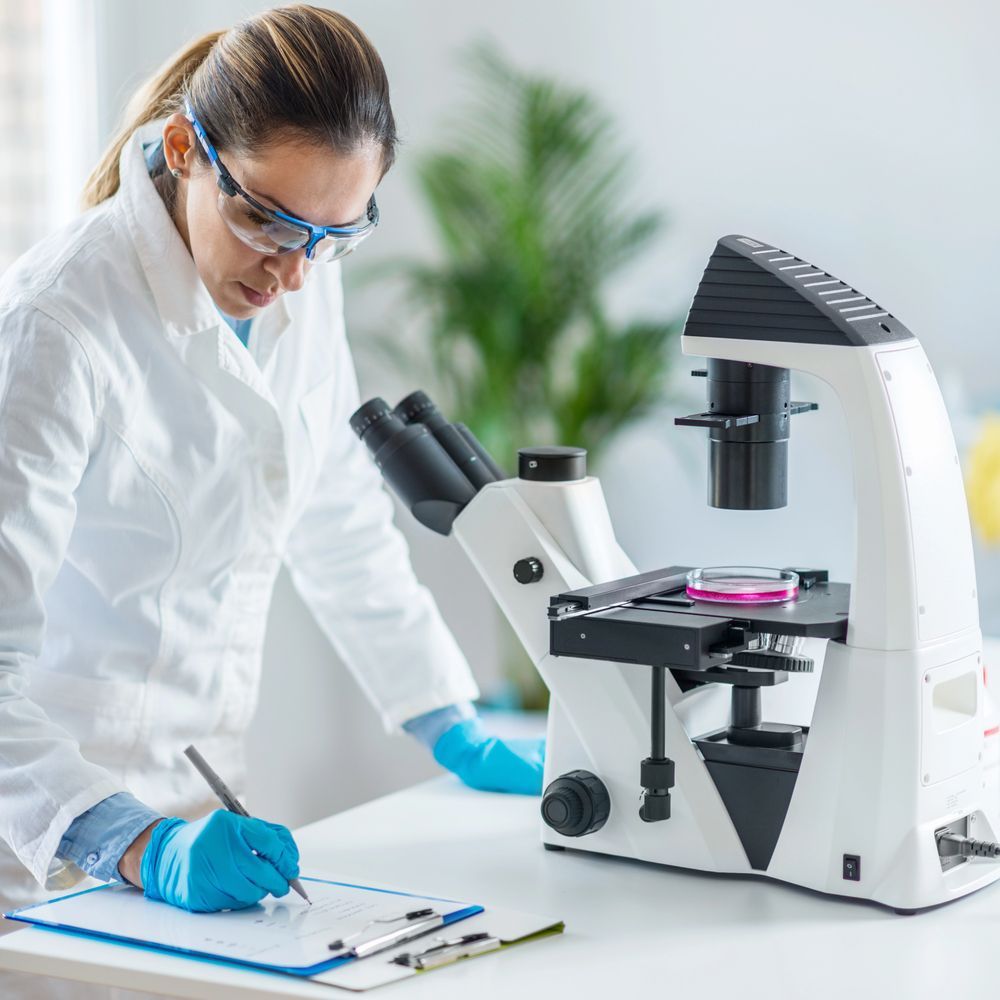 Un científico con bata blanca y gafas de seguridad toma notas mientras observa una muestra en un microscopio de laboratorio.
