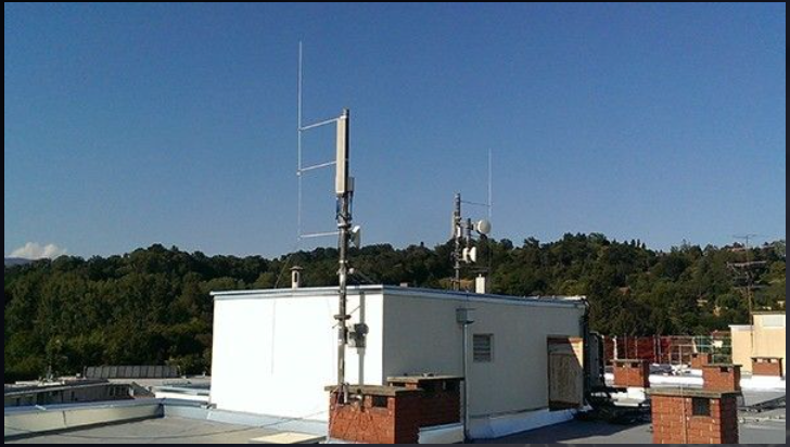 Ein Dach mit einer hohen Antennenanlage und einem weißen Technikunterstand vor klarem blauem Himmel und grüner Baumreihe.