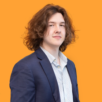 Young man with long brown hair in a blazer and patterned shirt against an orange background.