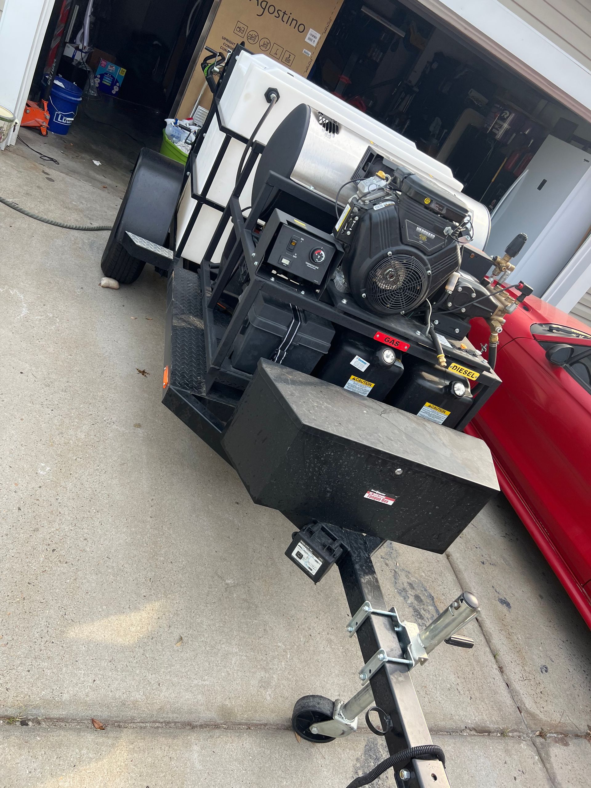 A pressure washing trailer with a black frame, white tank, black engine, and battery compartment; parked on concrete next to a red car.