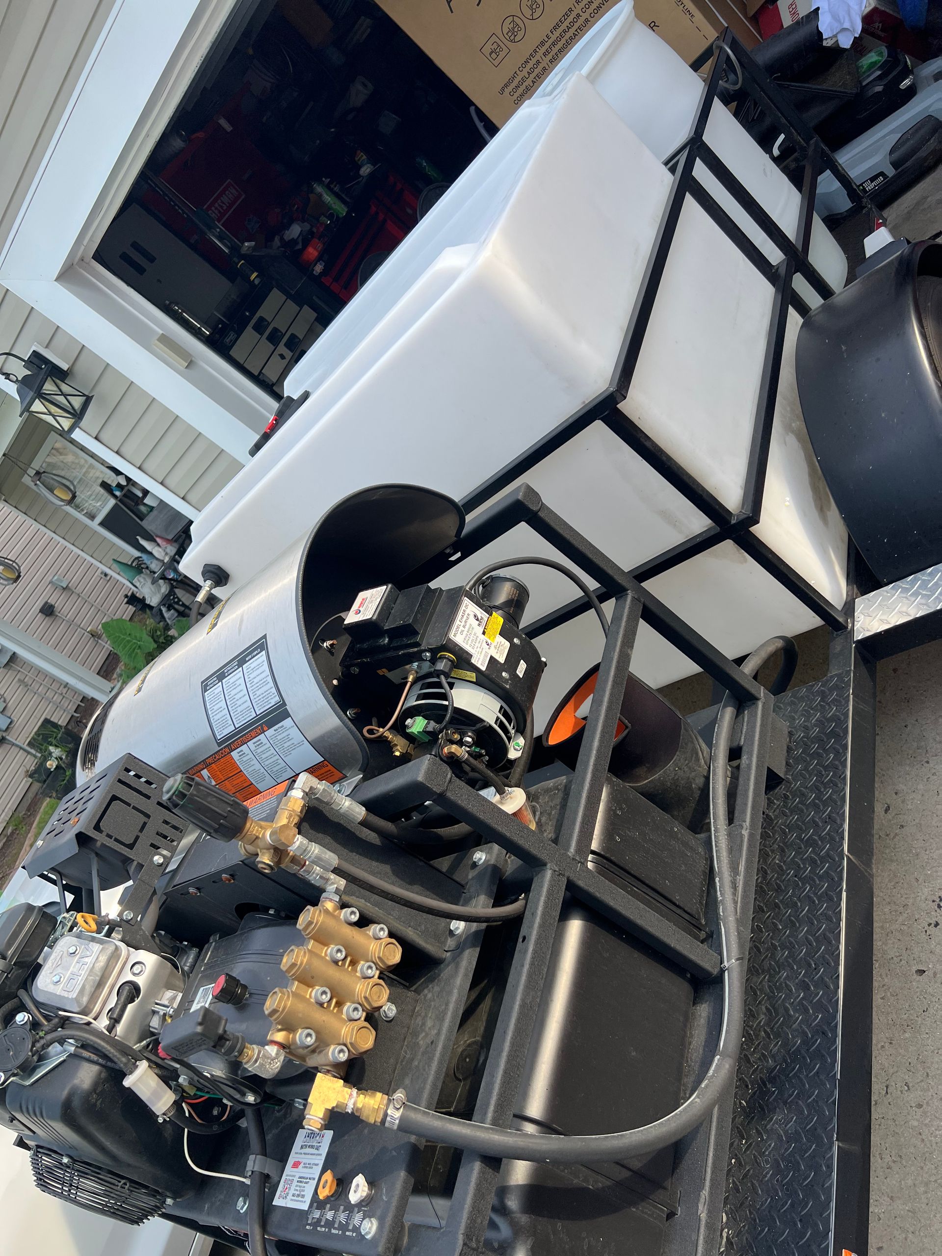 Pressure washer equipment mounted on a trailer with a water tank. The machinery includes a pump, engine, and hose connections.