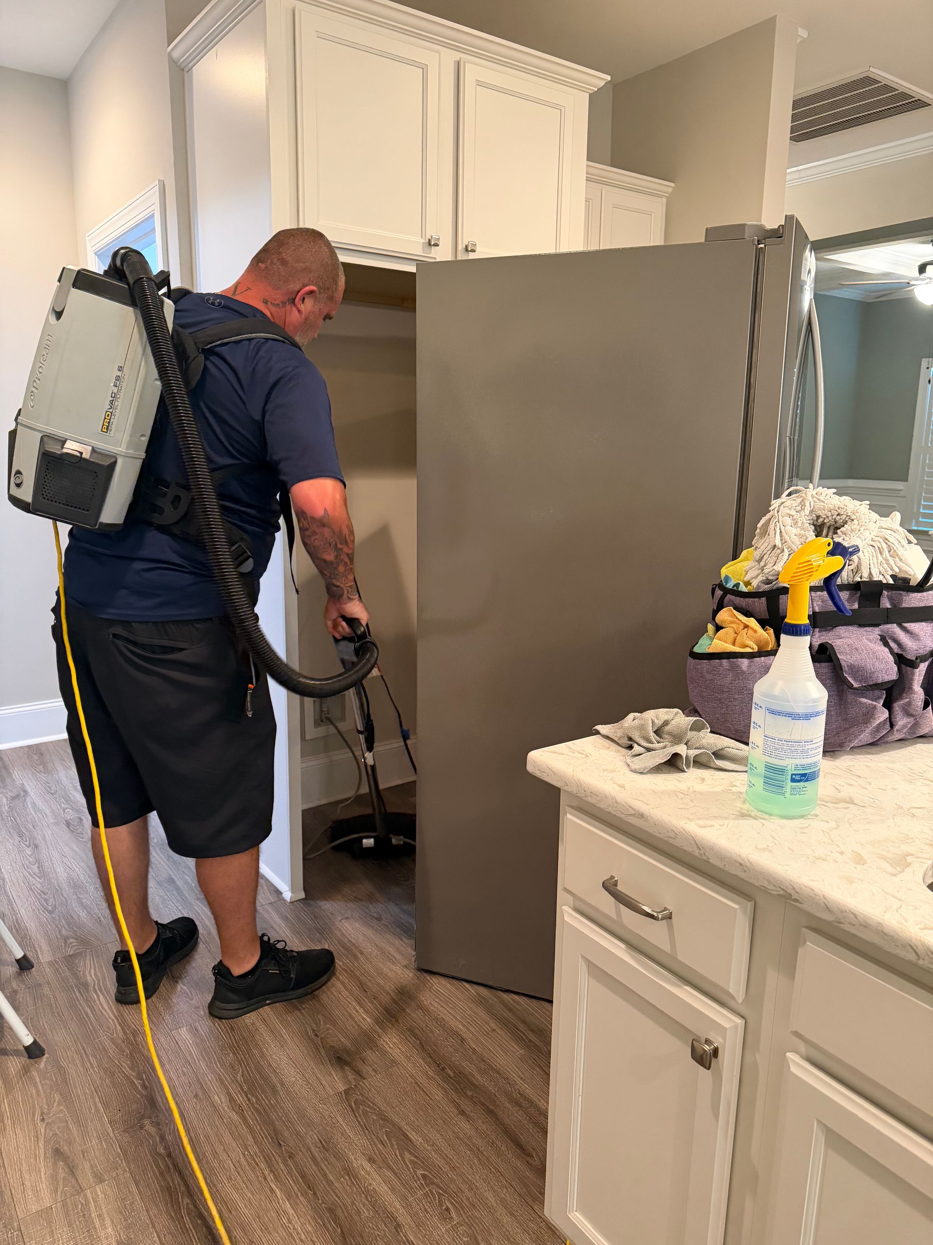 Person vacuuming kitchen floor beside gray refrigerator and white cabinets, near a counter with flowers and a bottle