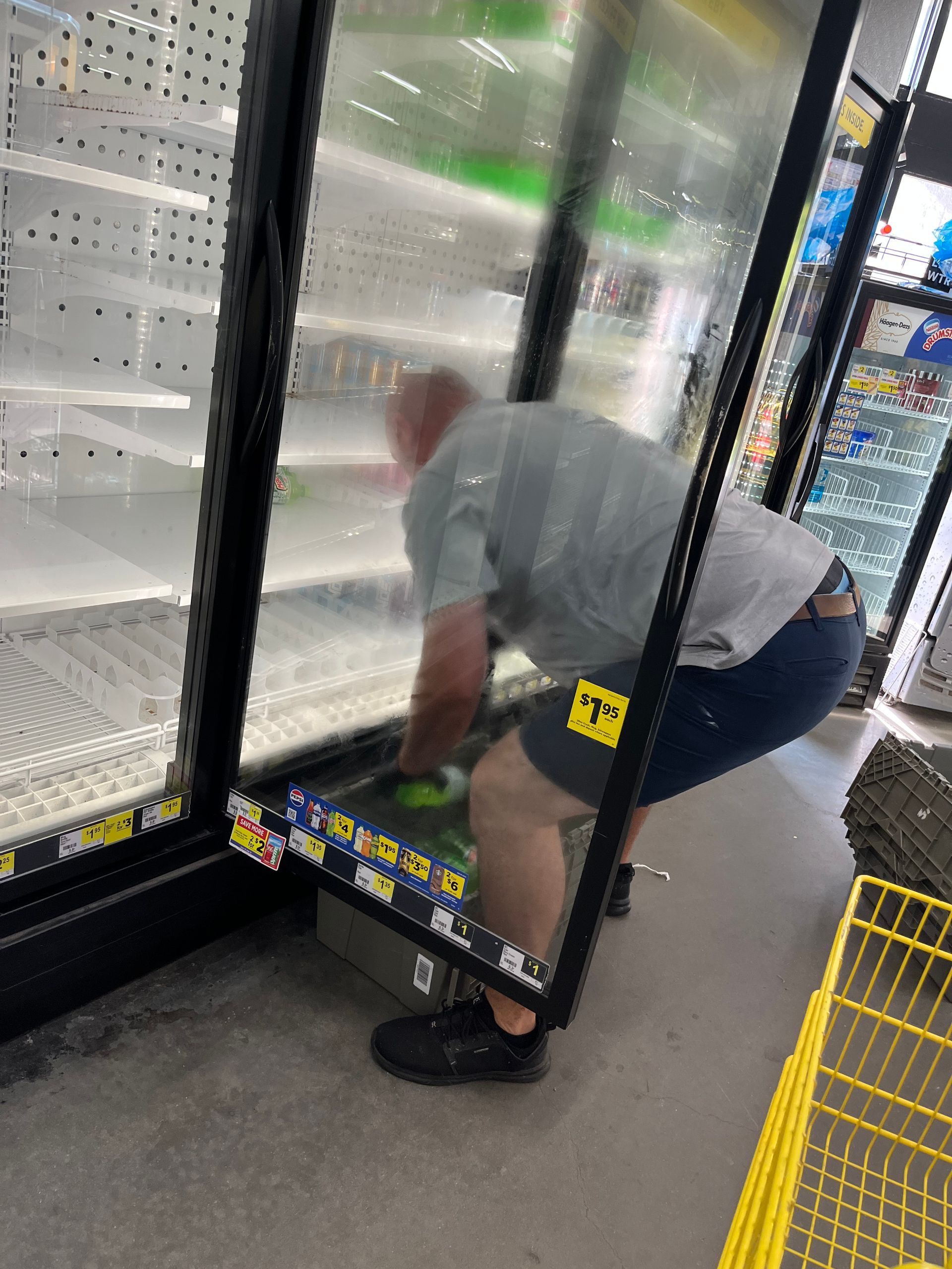 A man is reaching into a mostly empty refrigerated display case in a store. The case's glass door is open.