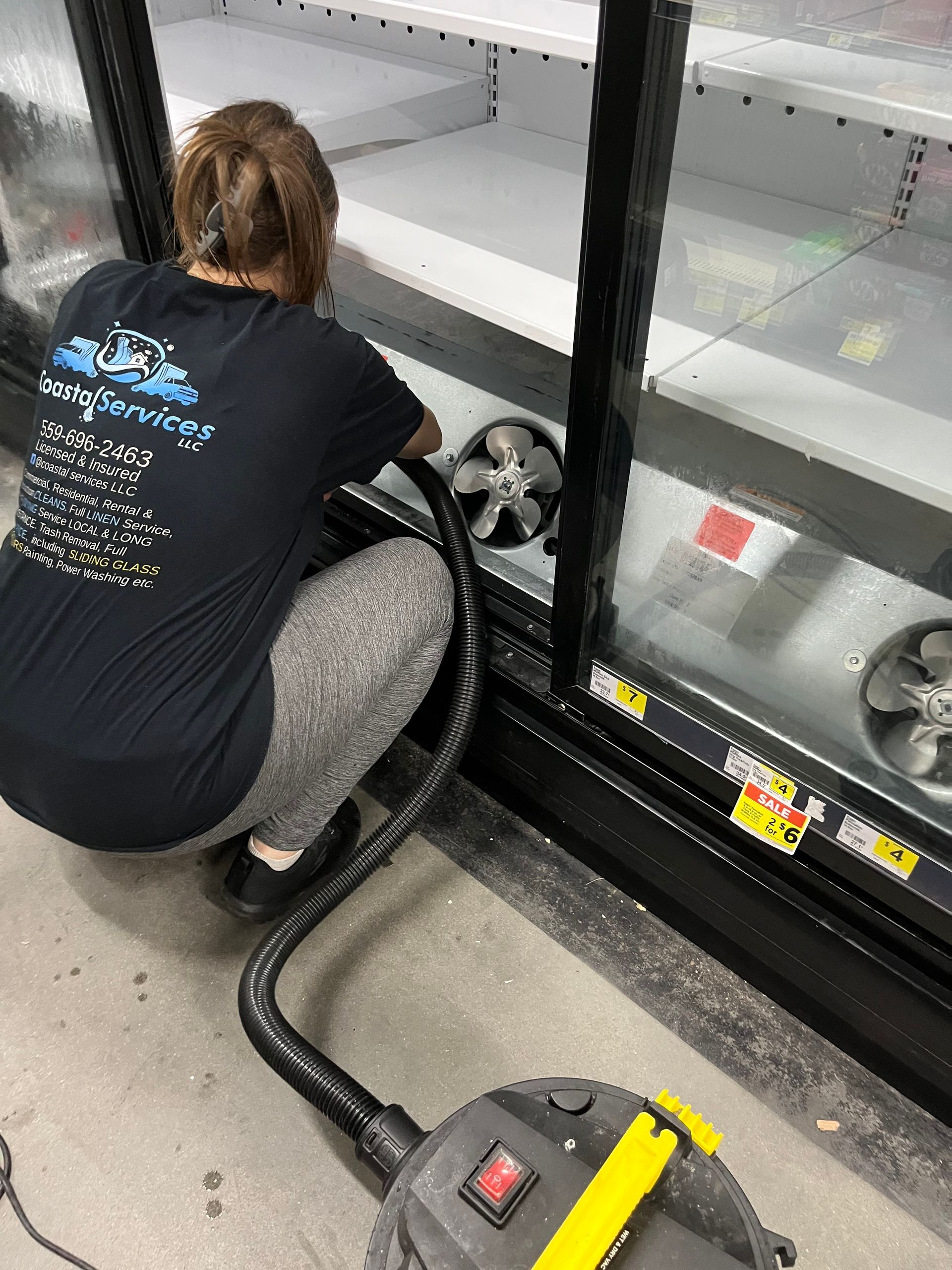 A person vacuums inside a commercial refrigerator. They wear a black t-shirt and gray leggings and kneel near the machine's fan.