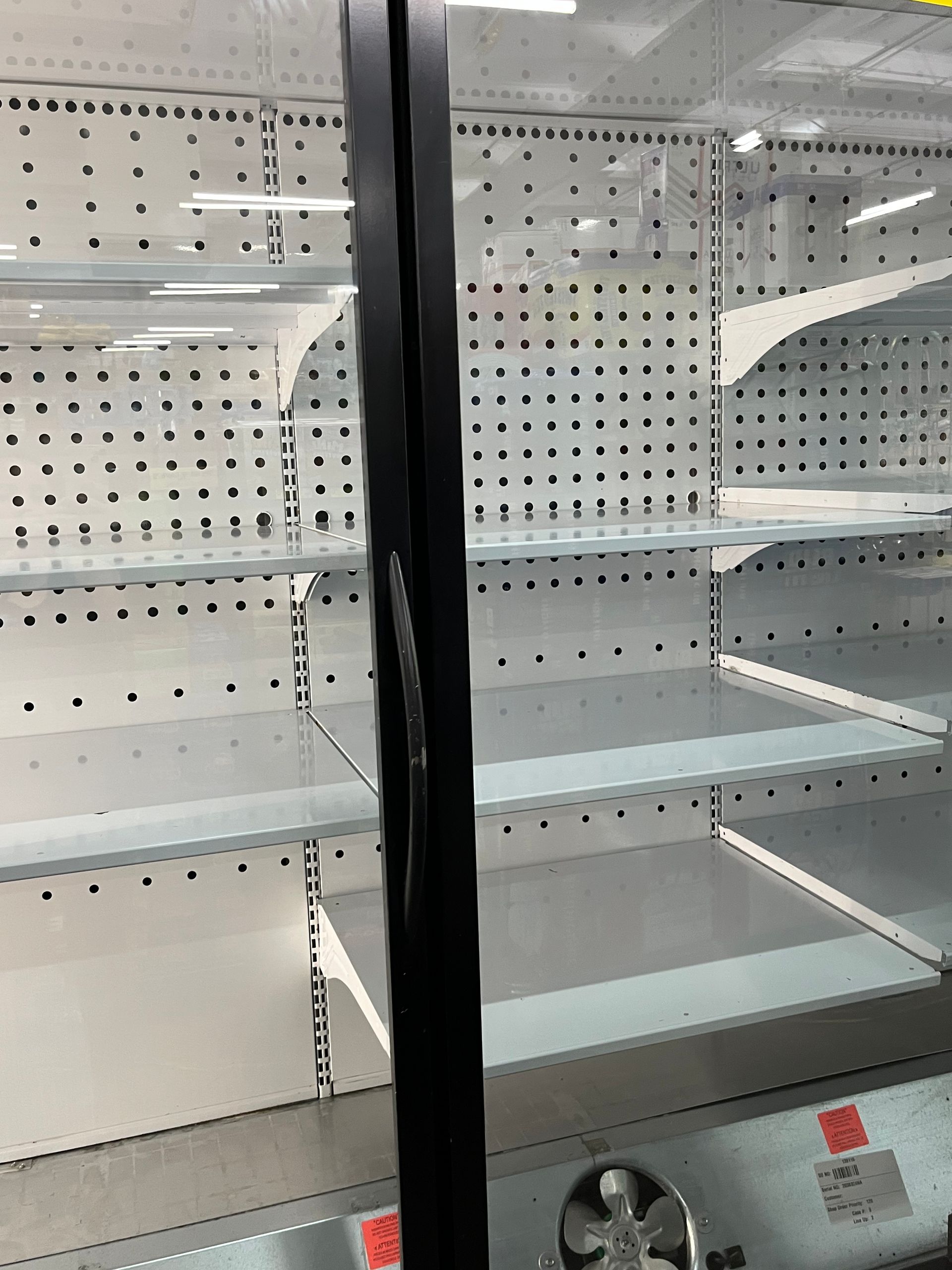 Empty refrigerated shelves in a store, likely due to supply chain issues. The shelves are white, the frame is black, and the setting is indoors.