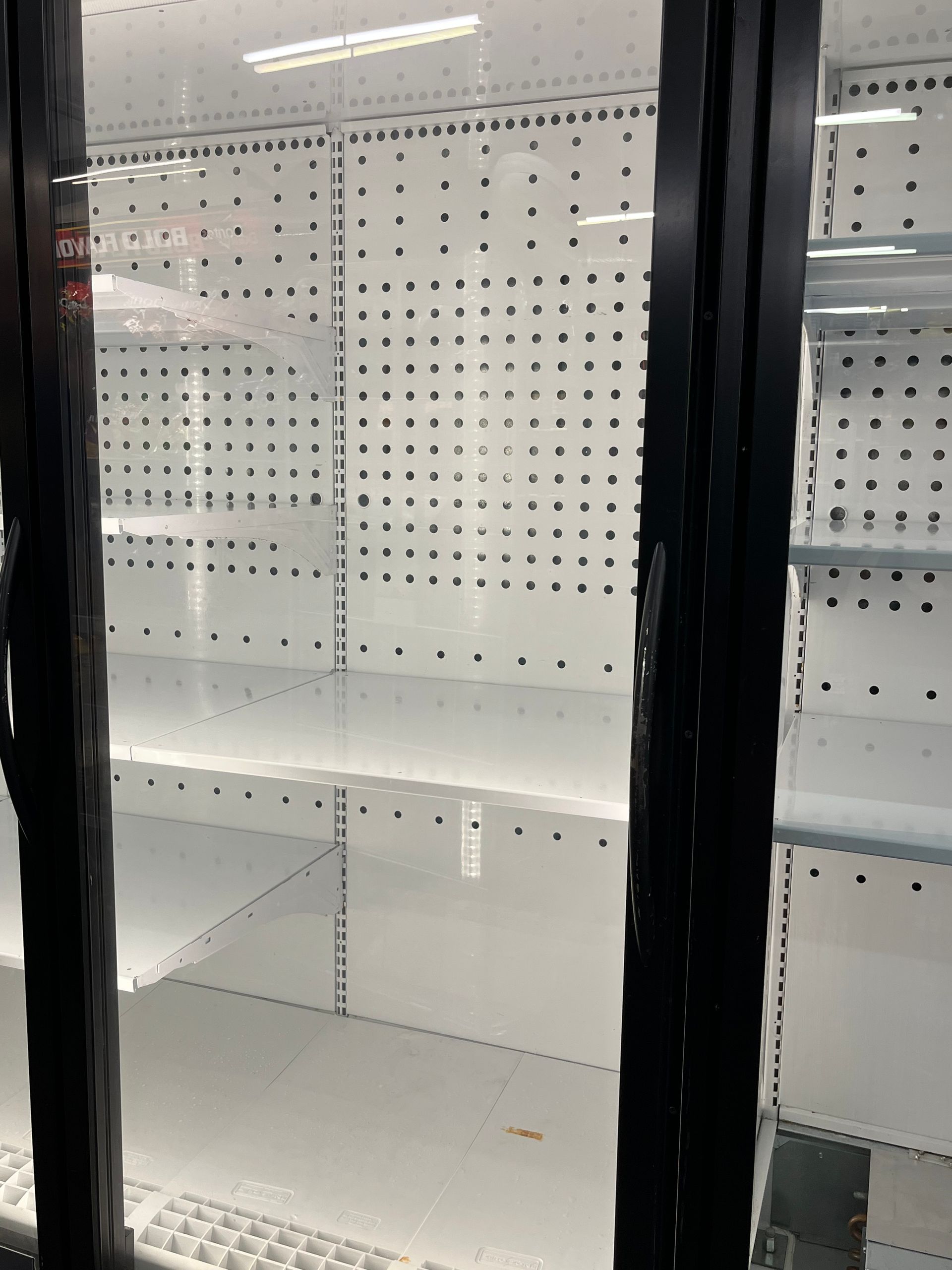 Empty refrigerator shelves inside a commercial cooler, viewed through glass doors.