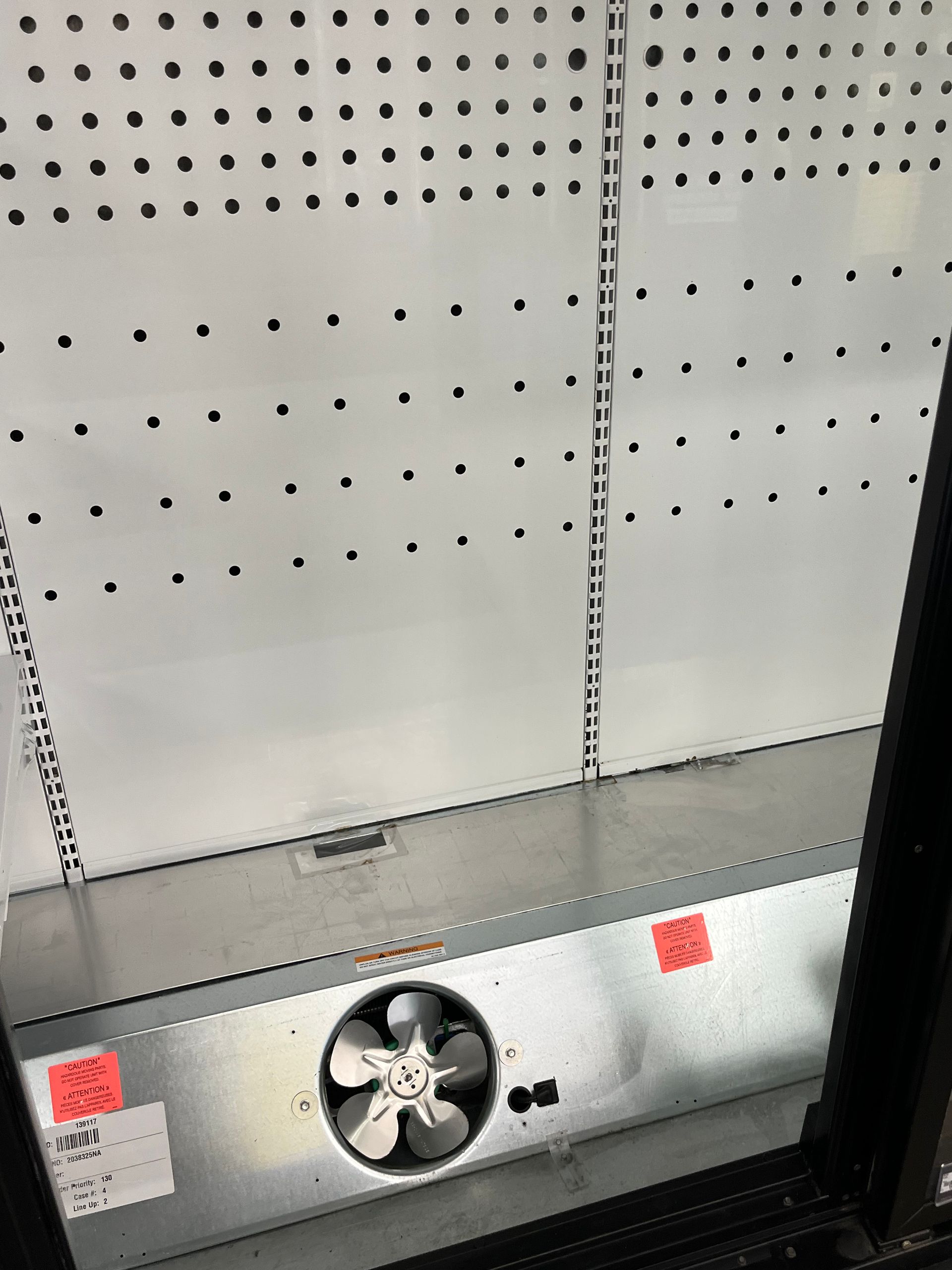 Interior view of a commercial refrigerator. A fan is visible, along with metal shelving supports and a metal back panel.