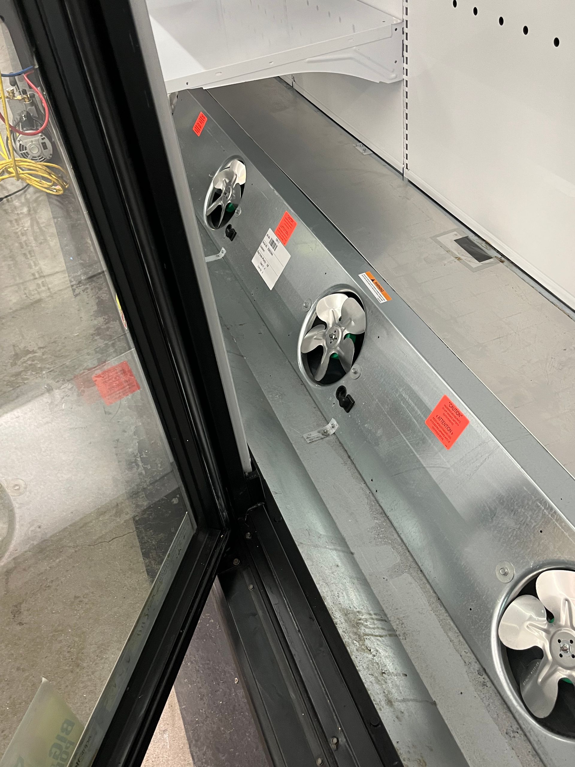 Interior of a commercial refrigerator with three visible fans and a partially open glass door, revealing metal surfaces and components.