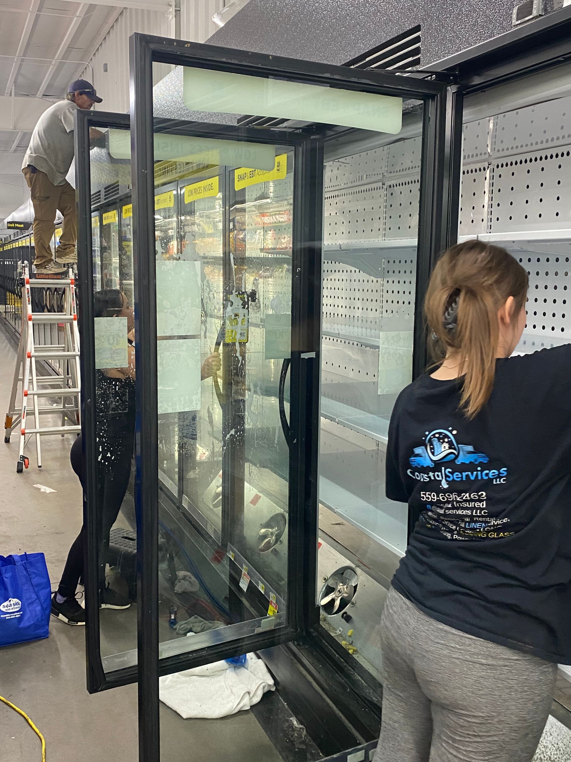 Three people installing a large refrigerated display case inside a store. One person stands on a ladder, while others work inside the unit.