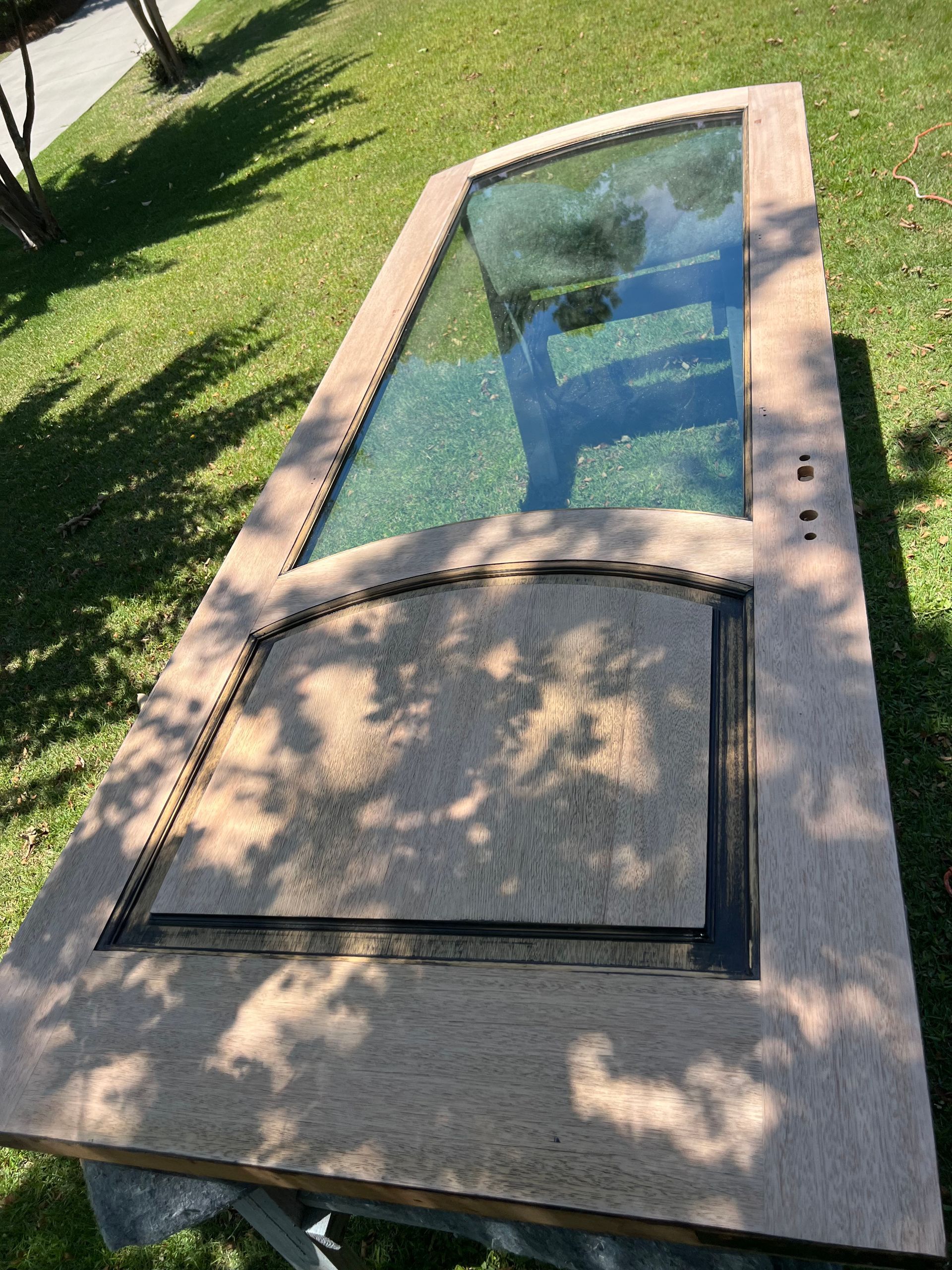 A wooden door with a large glass window panel, lying on grass outdoors in sunlight. The bottom half has a textured panel.