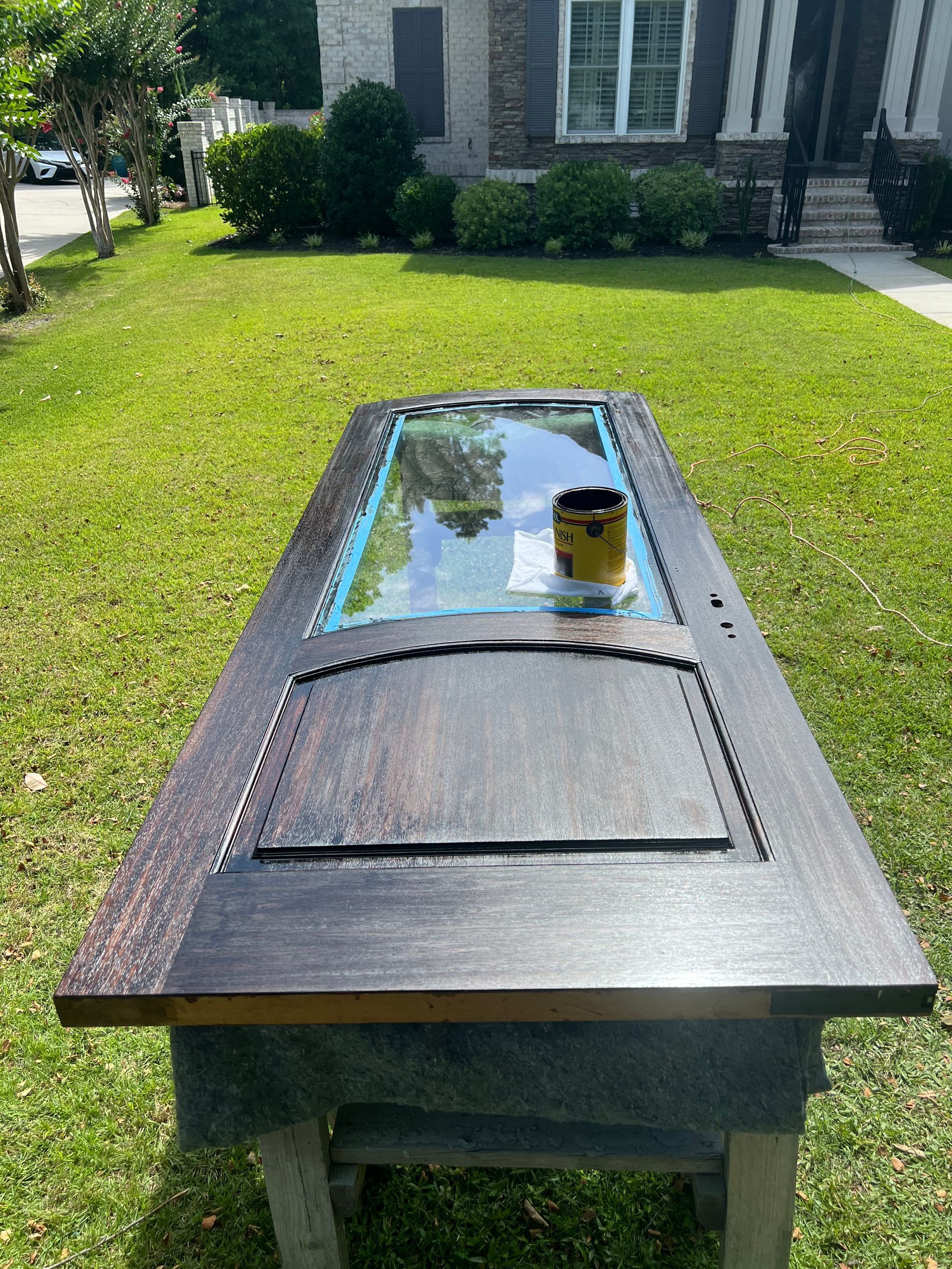 A dark-stained door repurposed as a table with a glass insert, resting on a makeshift stand, outdoors on a sunny day.