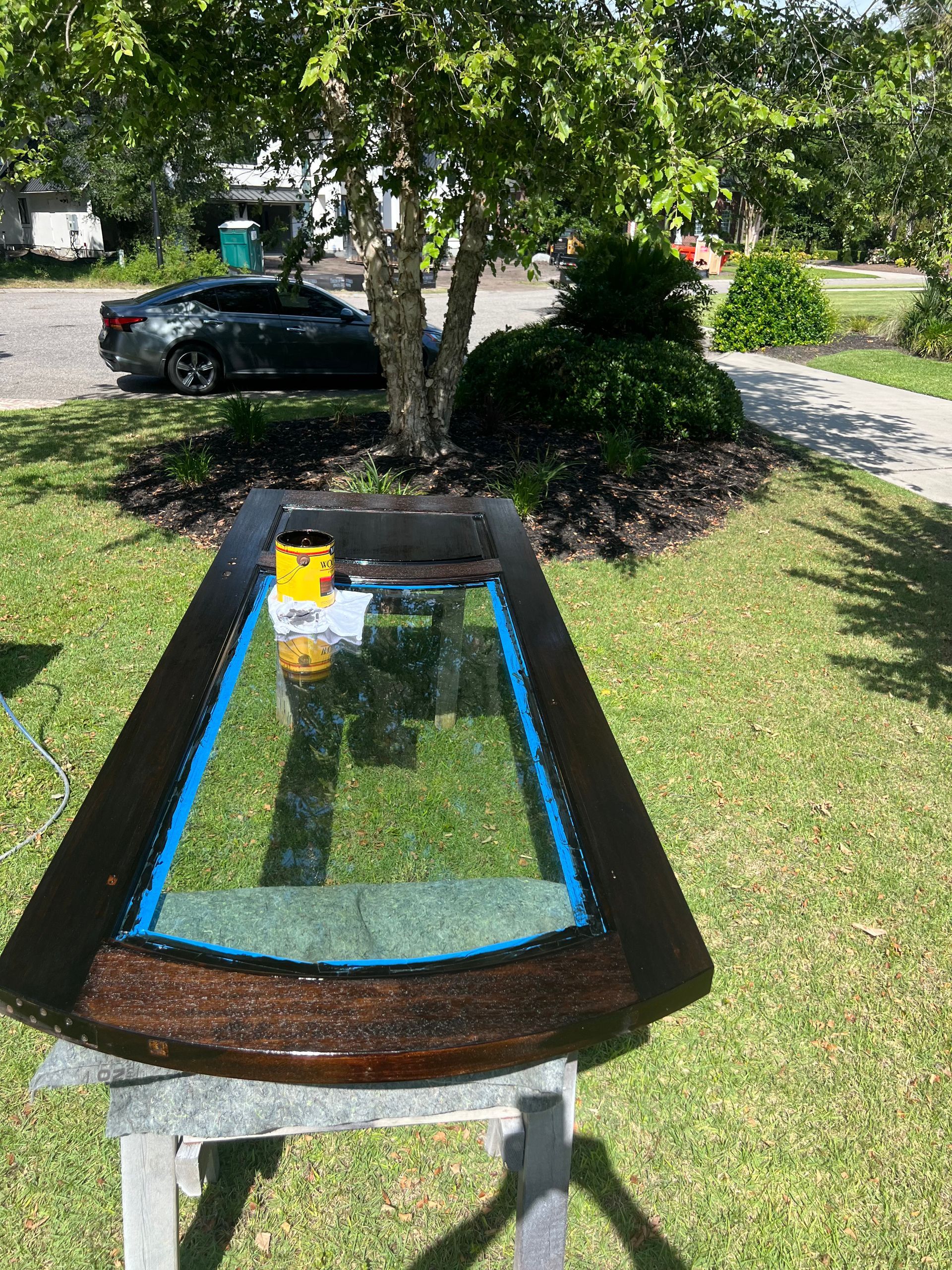 A wooden door with glass panes sits on a sawhorse outside. A yellow container and blue tape are on the door. The background includes grass, a car, and a tree.