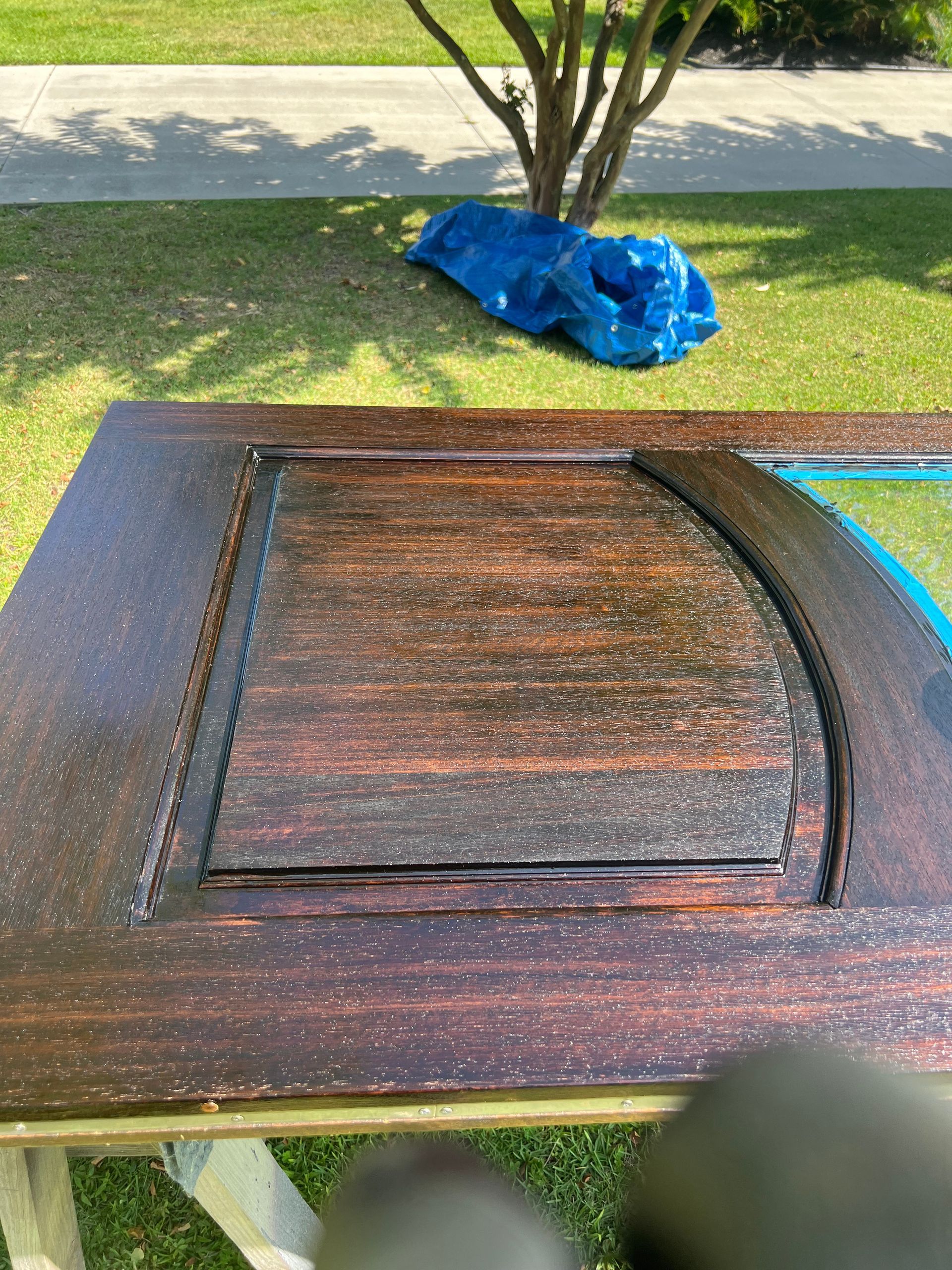 A freshly stained, dark wooden door laid on sawhorses outdoors. The door has a rectangular inset with a curved top. Blue tarp in the background.