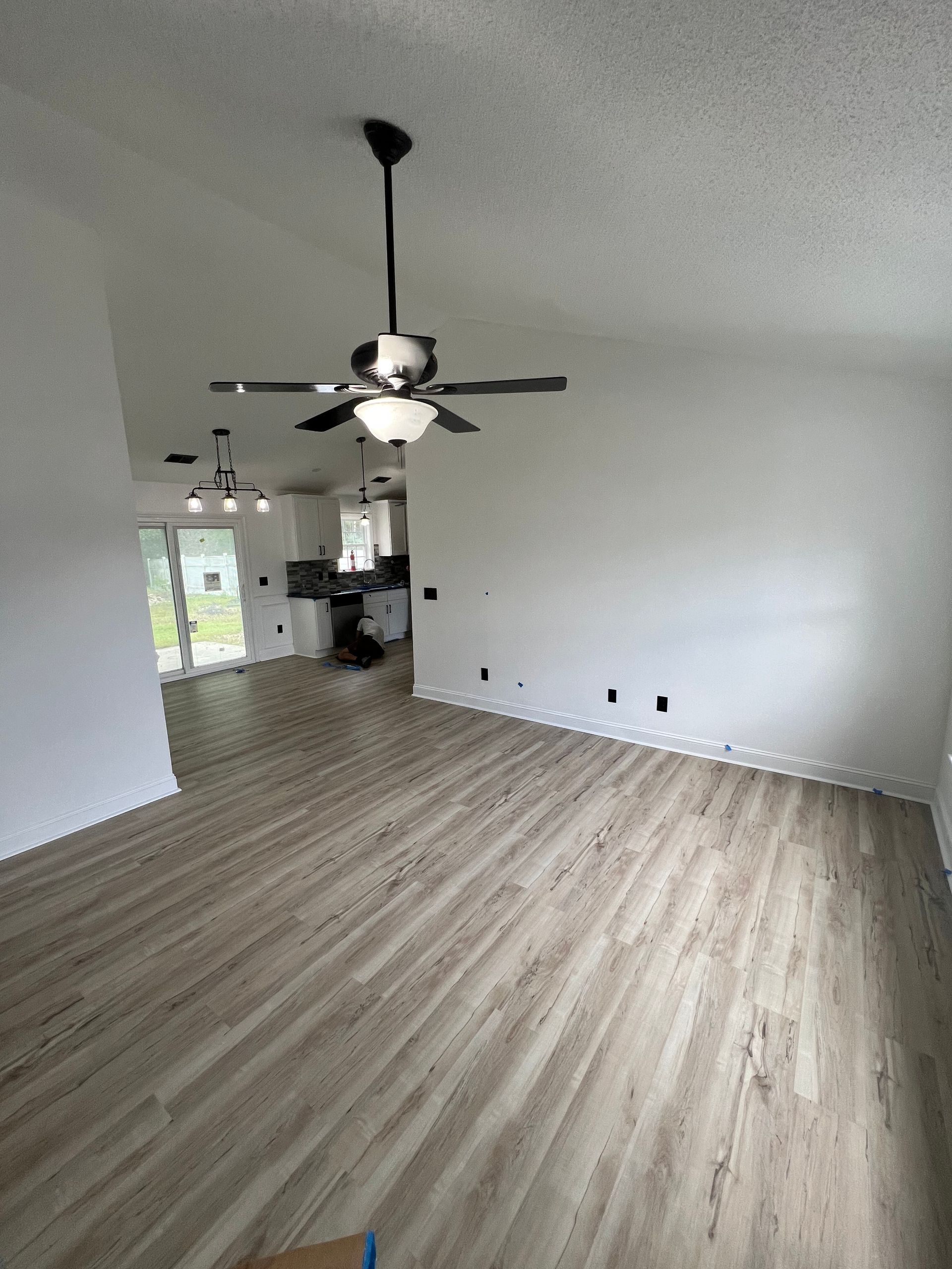 An empty living room with wood-look flooring, white walls, and a black ceiling fan.  The room has a vaulted ceiling and opens into a kitchen area.