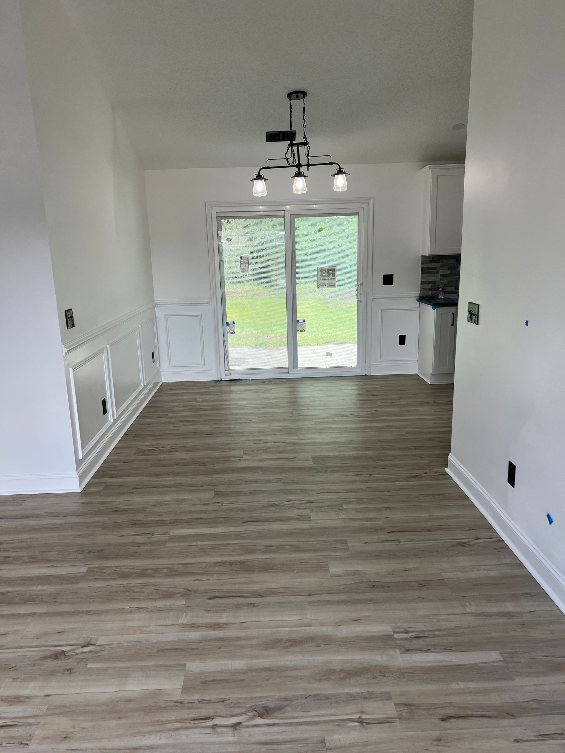 Empty dining room with wood-look flooring, white walls, and a sliding glass door to a green outdoor view. A dark chandelier hangs above.