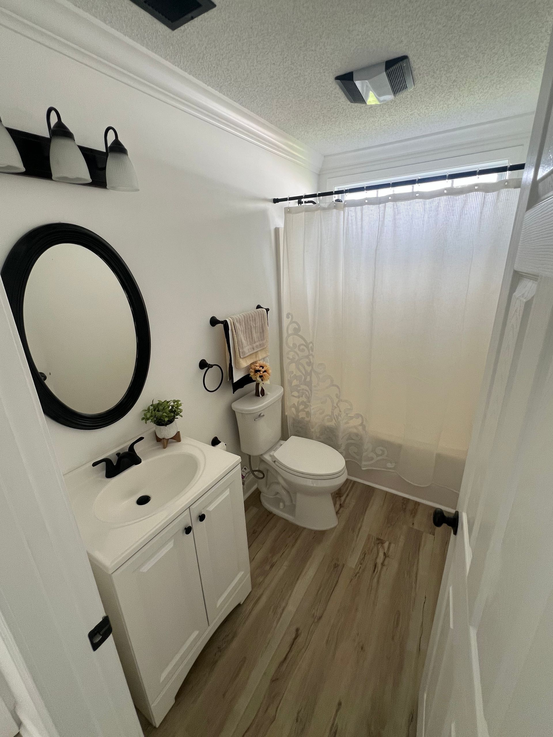 A small, modern bathroom with white walls, a white vanity, and a toilet. Black fixtures and a light wood-look floor contrast the white.