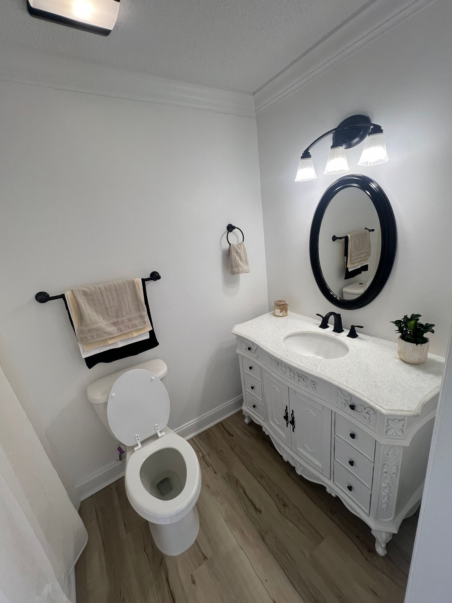 A small bathroom with a white vanity, toilet, and walls. Includes a black-framed mirror, towel racks, and faux wood flooring.