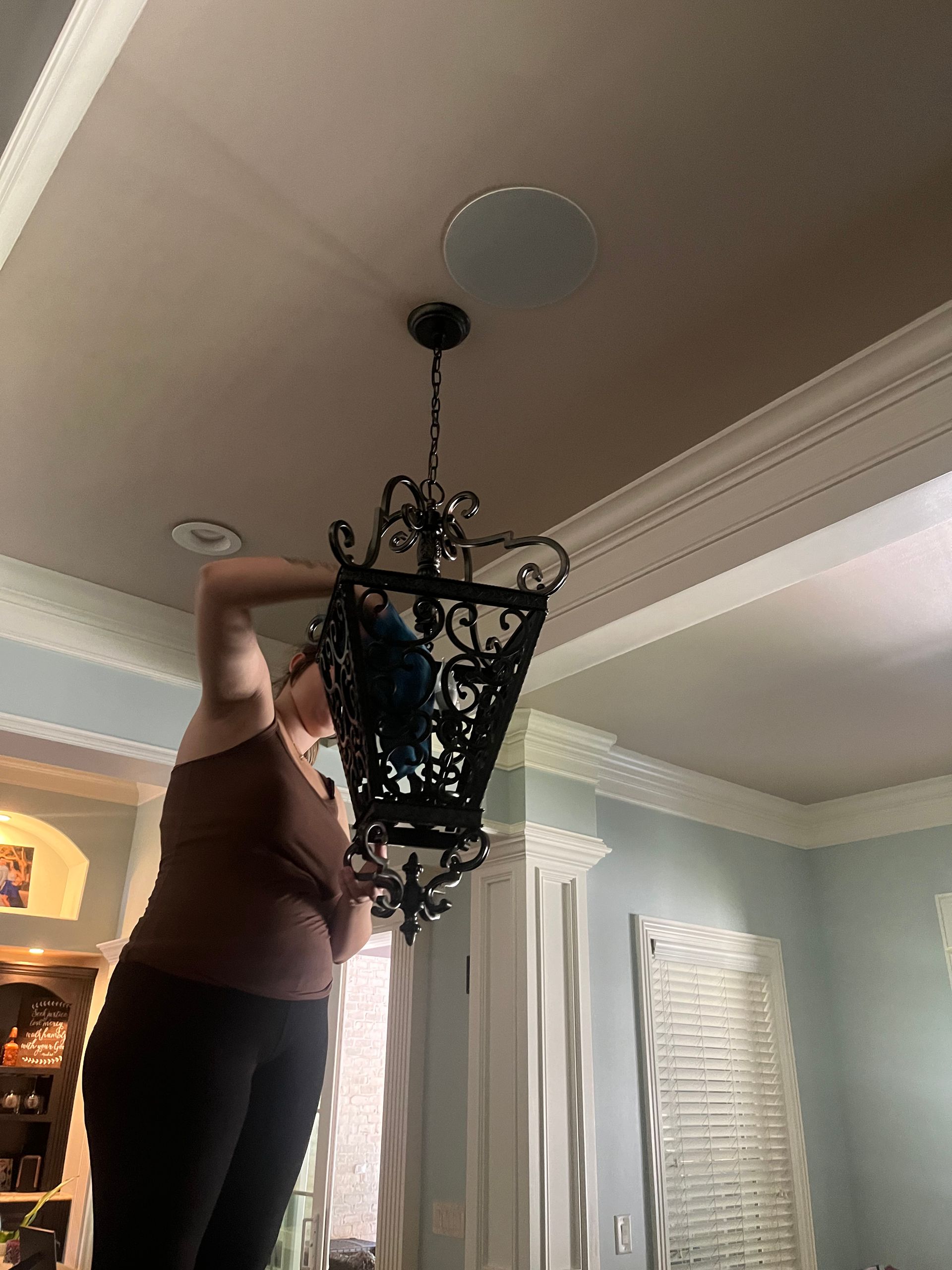 A person on a stepladder adjusts a black, ornate chandelier hanging from a light-colored ceiling. The room has pale blue walls and white trim.