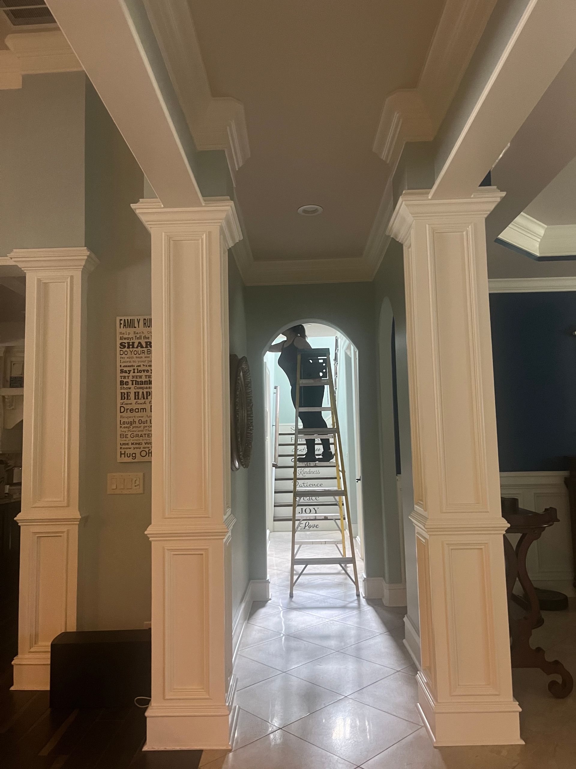 A hallway with white pillars and a person on a ladder. Light blue walls and patterned floors.