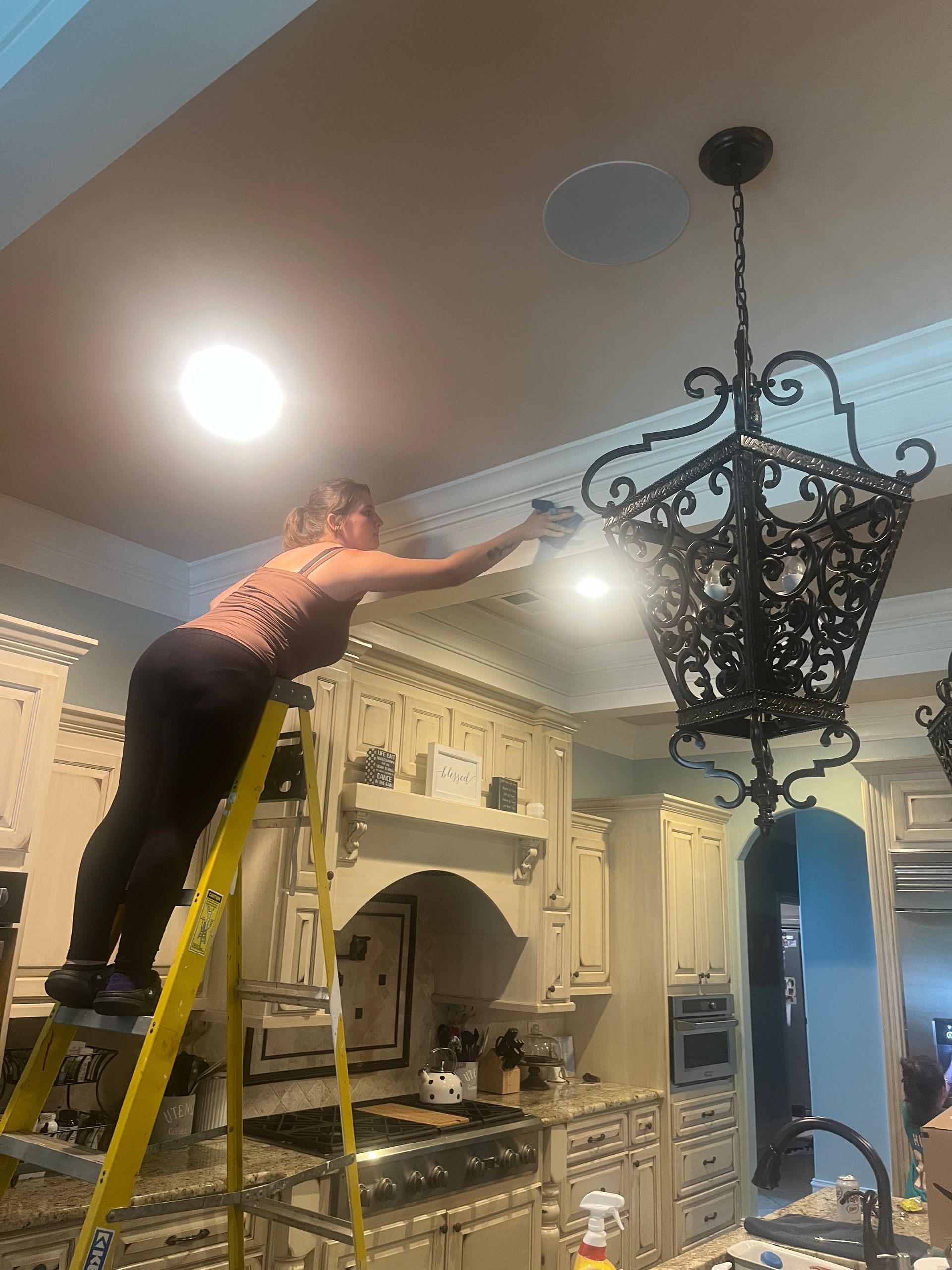 Woman on a ladder reaching toward the ceiling in a kitchen. She's near a chandelier and recessed lights, with ornate cabinets below.