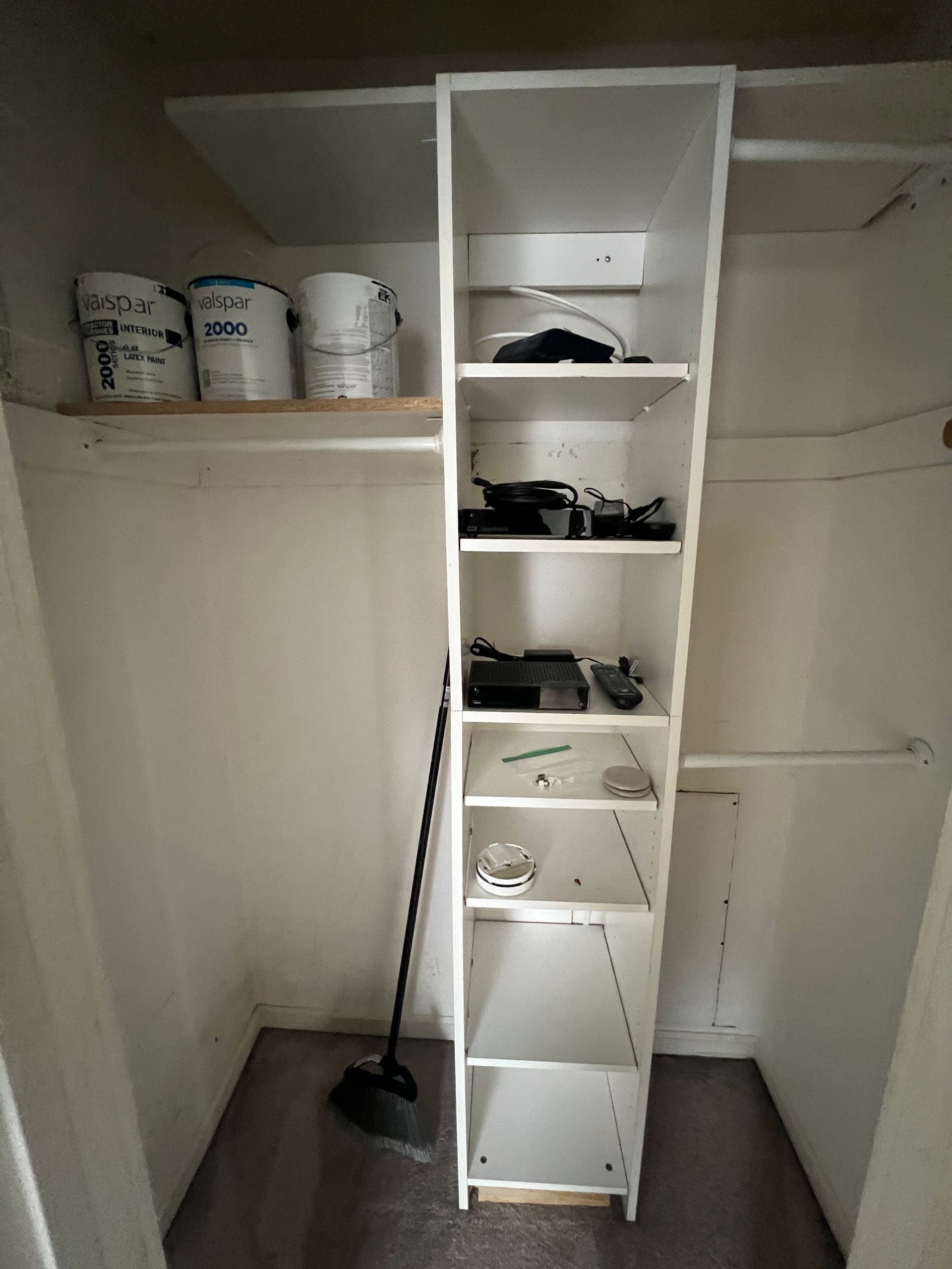 A narrow white shelf unit inside a closet, holding various objects. Paint cans sit on a shelf above, and a broom leans nearby.