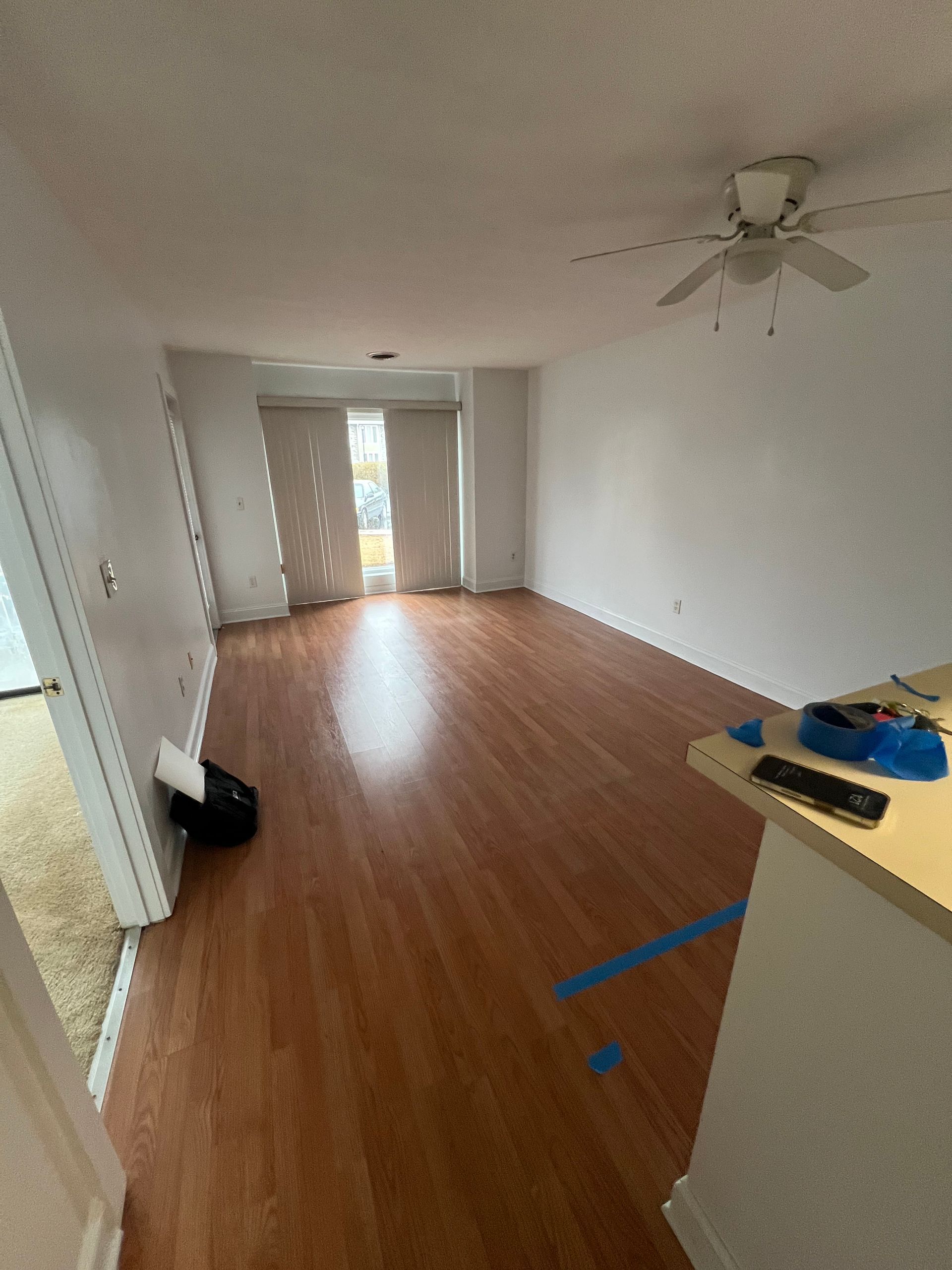 Empty living room with wood-look floors, white walls, and a sliding glass door. A ceiling fan is visible.