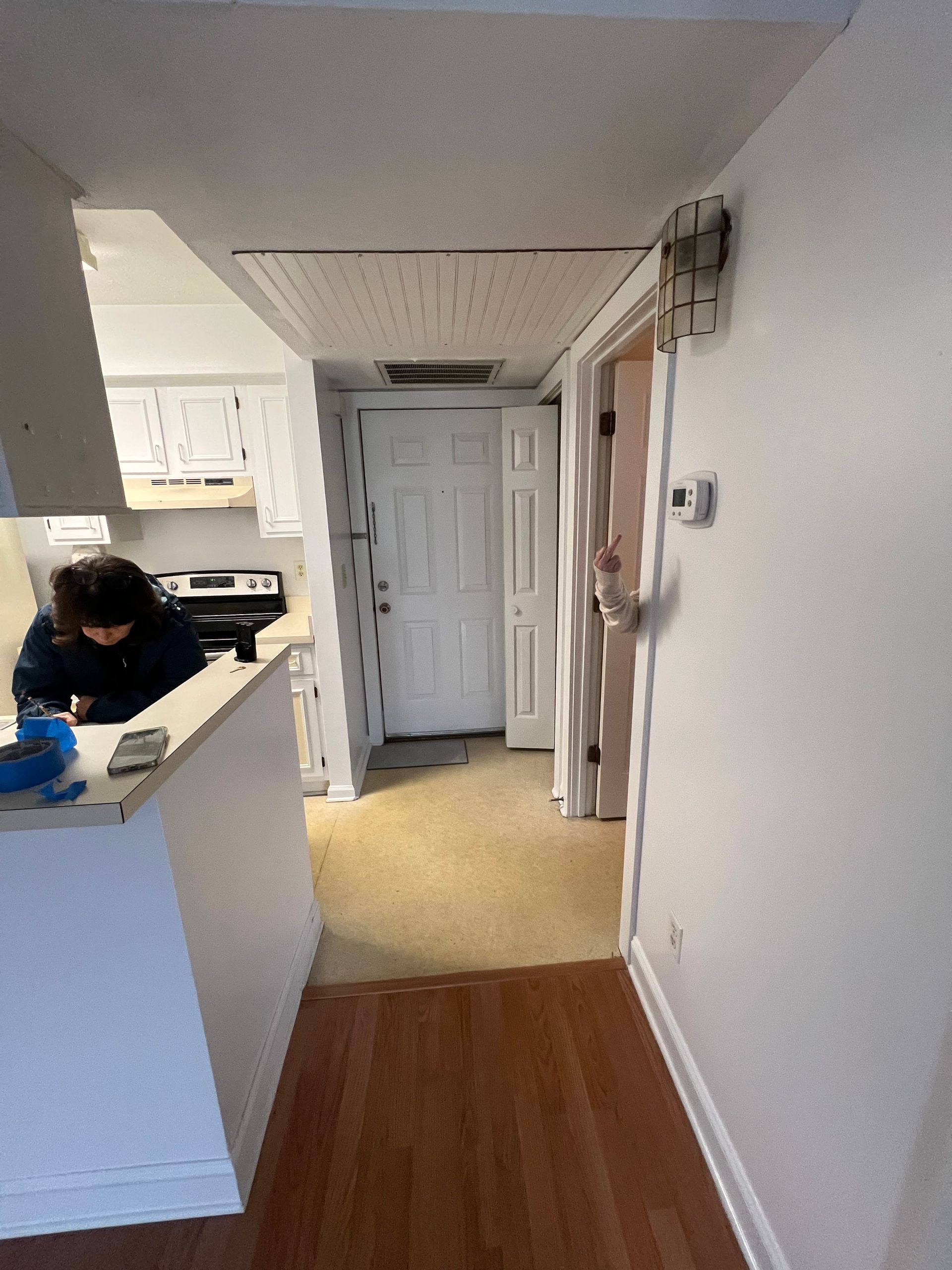 Hallway leading to a white door, with a kitchen and person on the left and a door on the right.