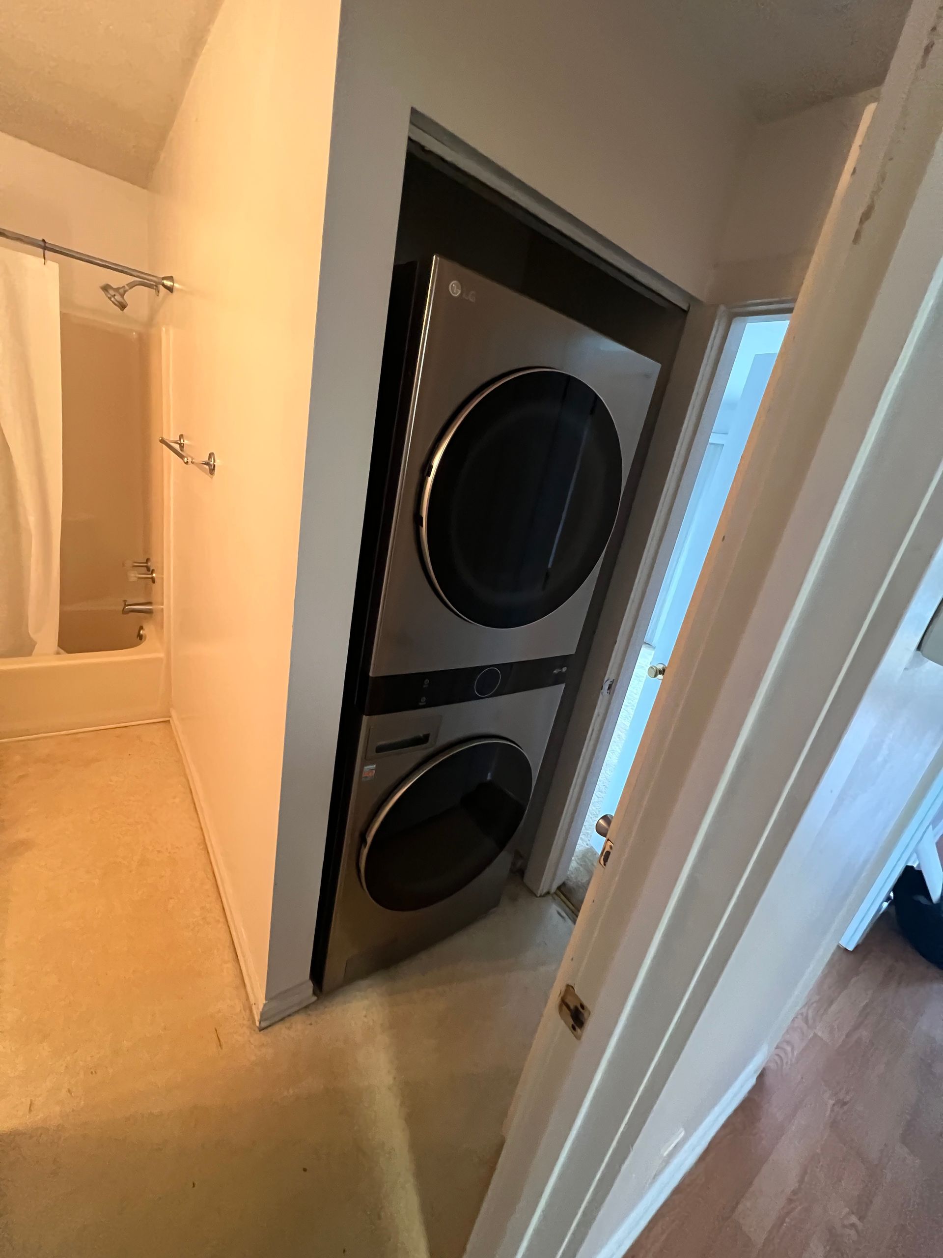 Stacked washer and dryer unit in a narrow closet next to a bathroom with a shower curtain. The machines are stainless steel.