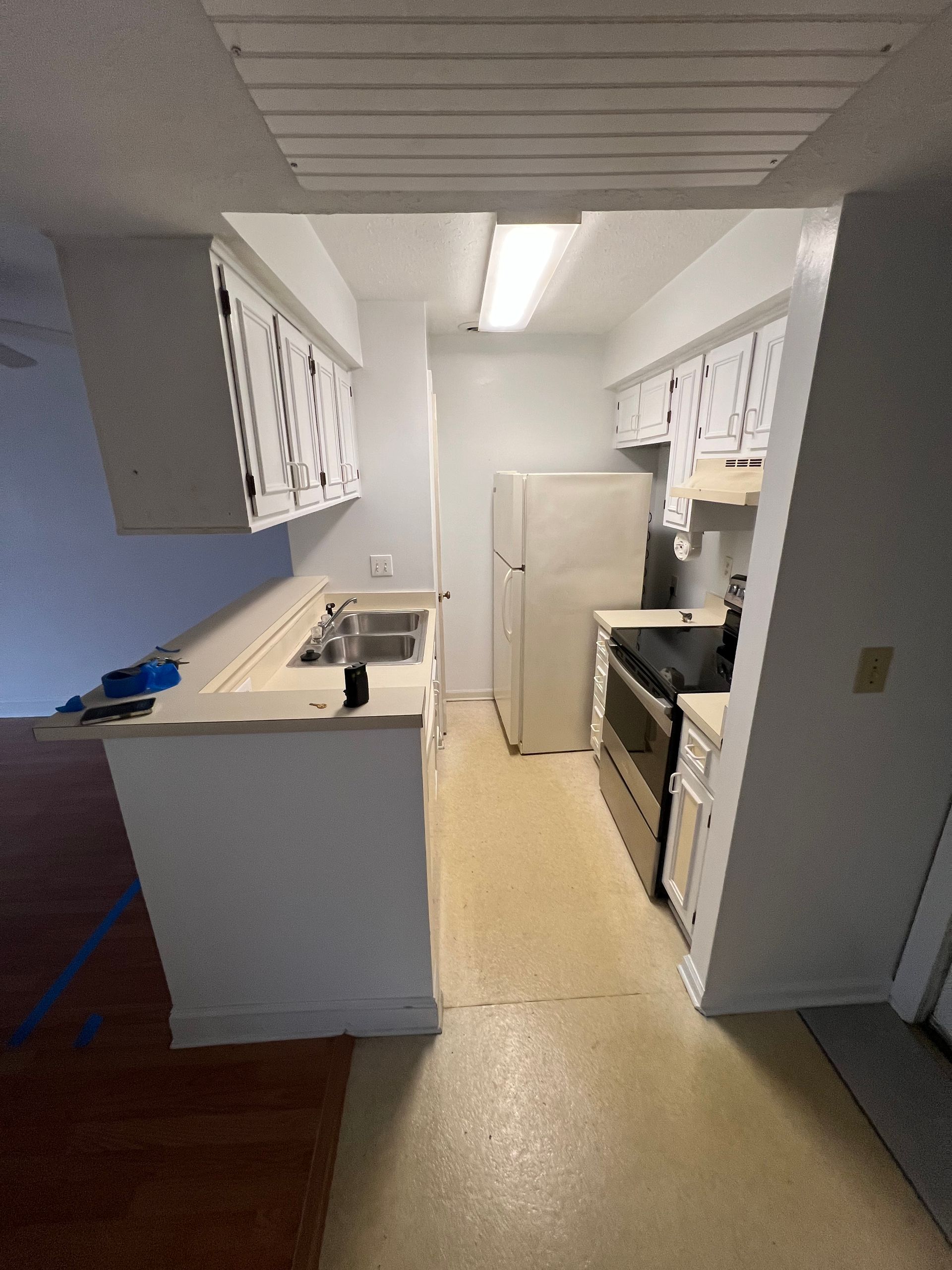 A small, narrow white kitchen with a refrigerator, stove, and sink. Cabinets line the walls, with a counter separating the kitchen from another room.