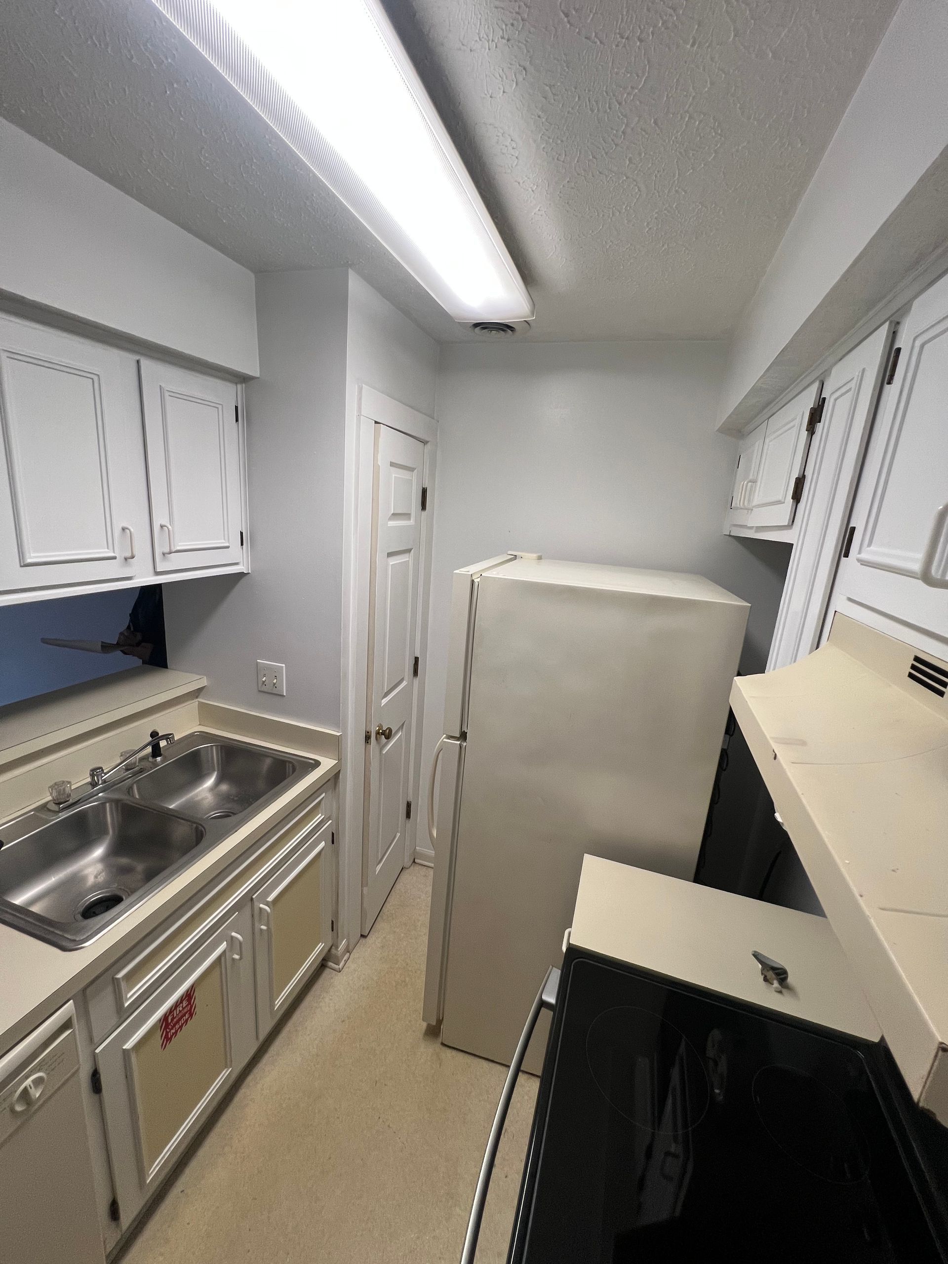 Narrow kitchen with white cabinets, a double sink, a refrigerator, and a stove. The walls and the cabinets are white and the floor is tan.