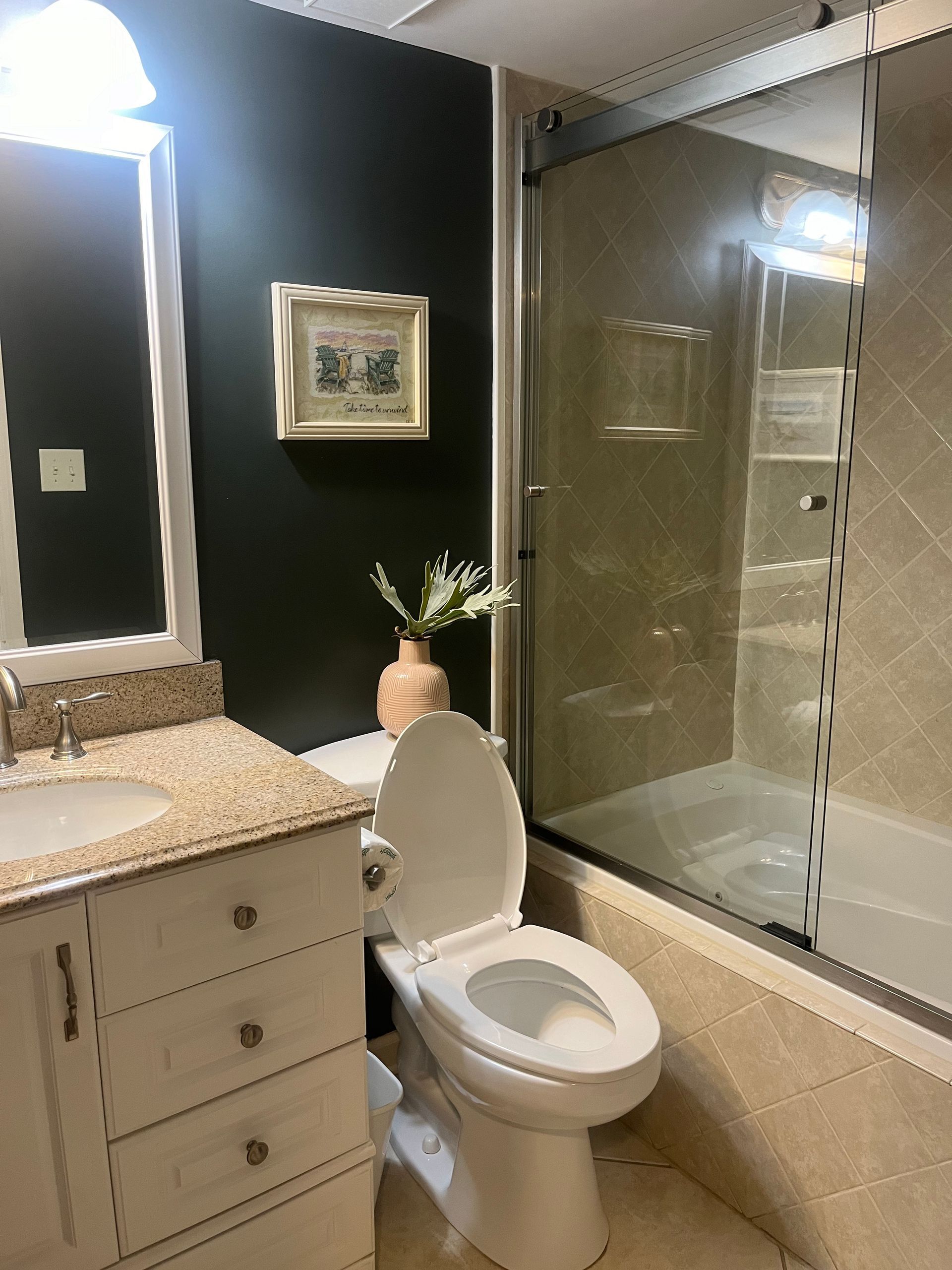Bathroom with dark wall, white vanity, toilet, and glass shower. A painting and vase with flowers sit on the wall.