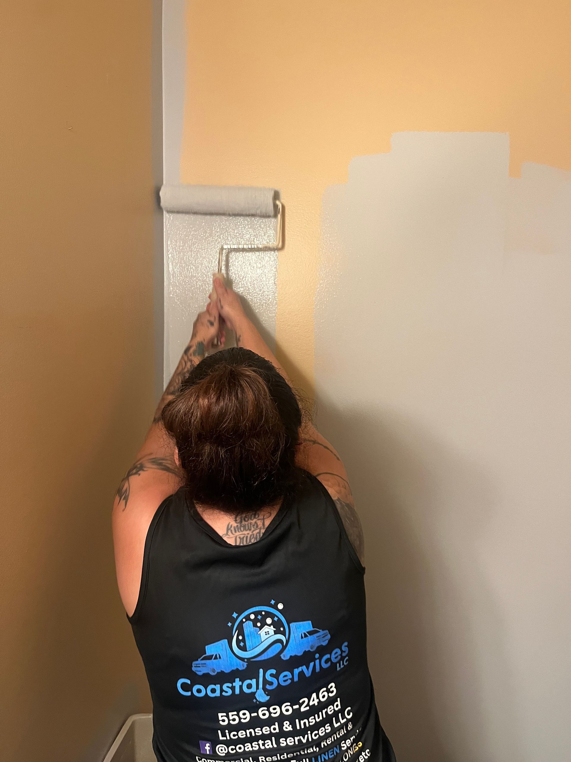 Person wearing a black tank top paints a wall using a roller. The wall is half yellow and half gray.
