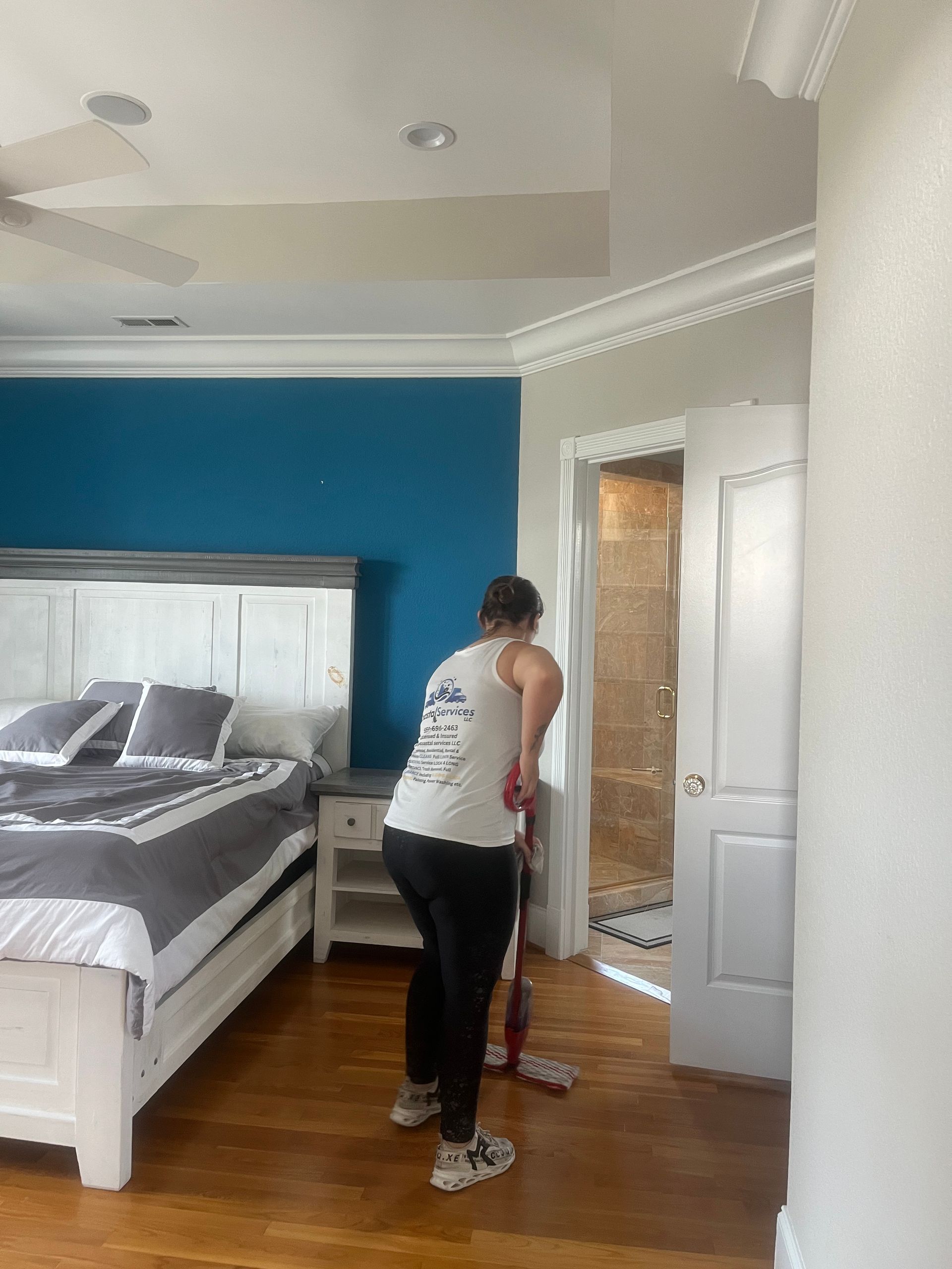Person mops a wooden floor in a bedroom with a blue accent wall.  A bed, nightstand, and open shower are visible.