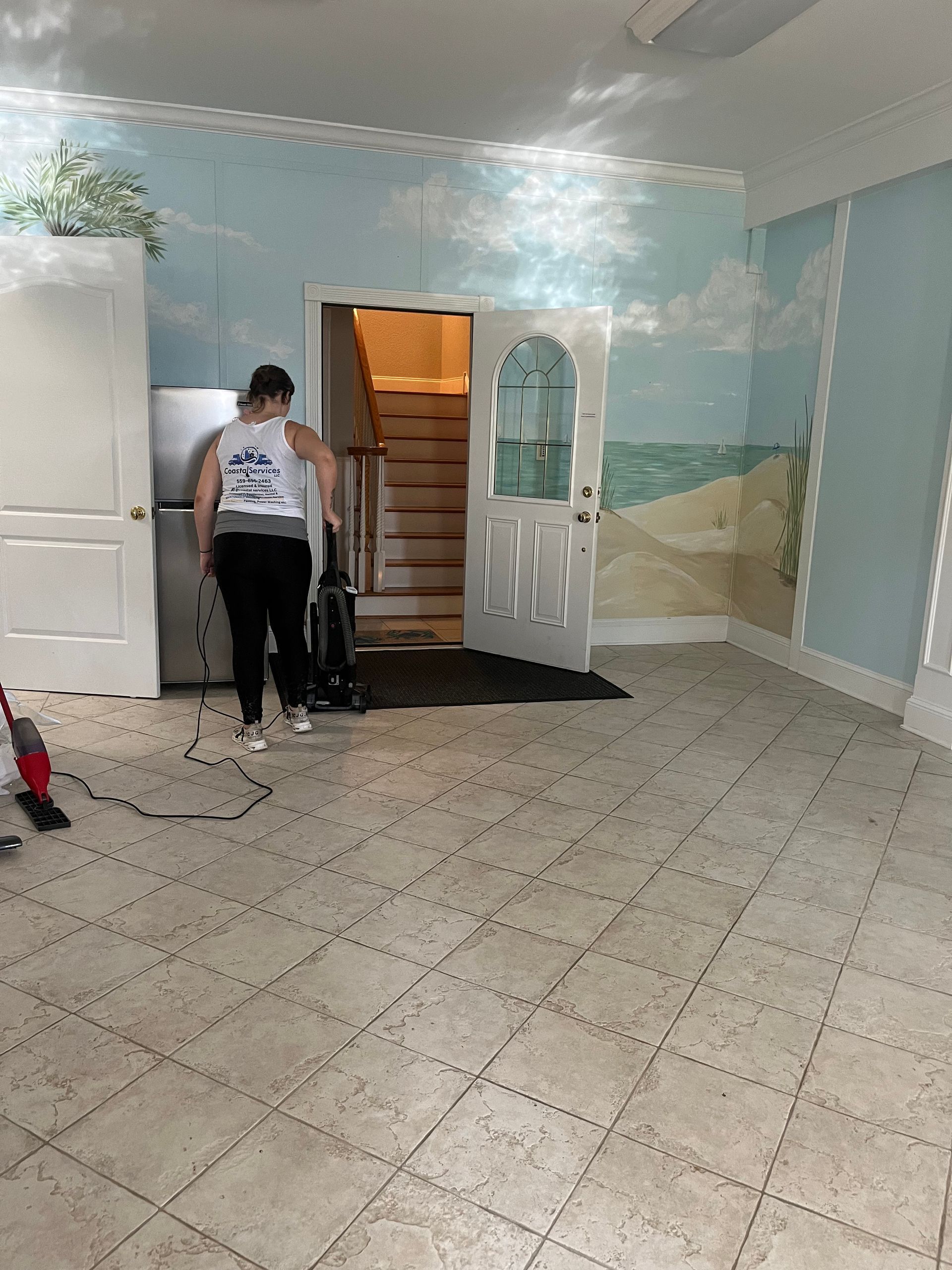 A person vacuums a room with a beach mural on the wall. They face away from the camera, and a doorway with stairs is in the background.