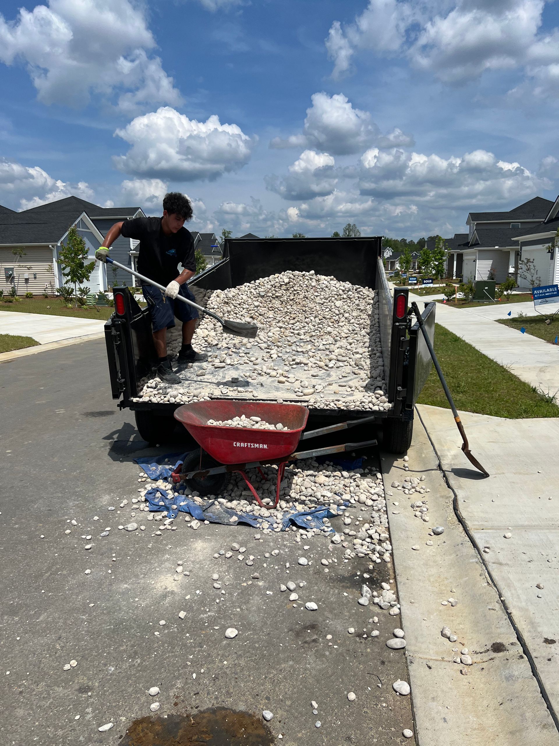 A person shovels gravel from a truck bed into a red wheelbarrow on a sunny residential street.