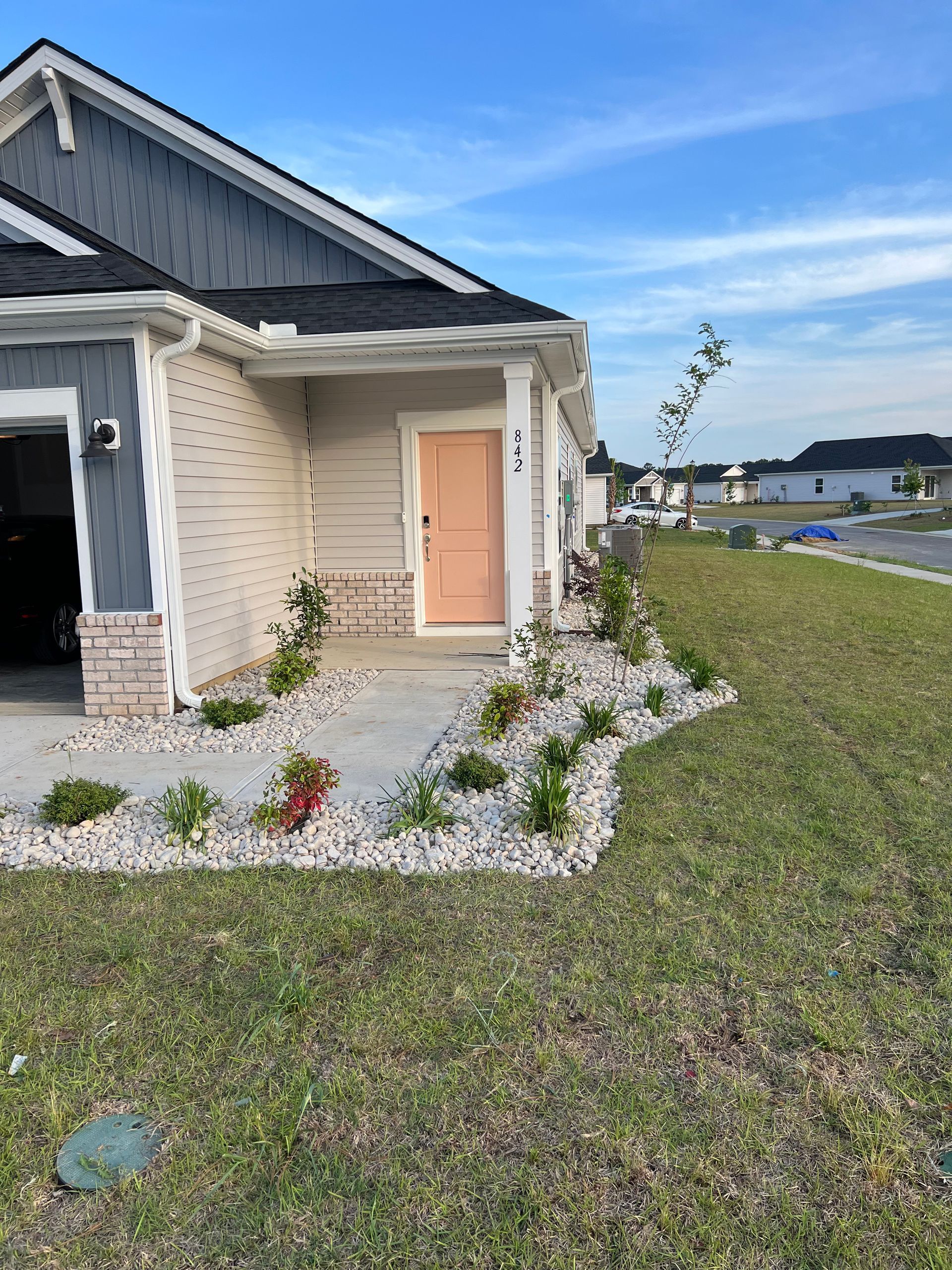Exterior view of a light-colored house with a peach-colored door. Landscaping includes gravel, grass, and small plants. Blue sky in the background.