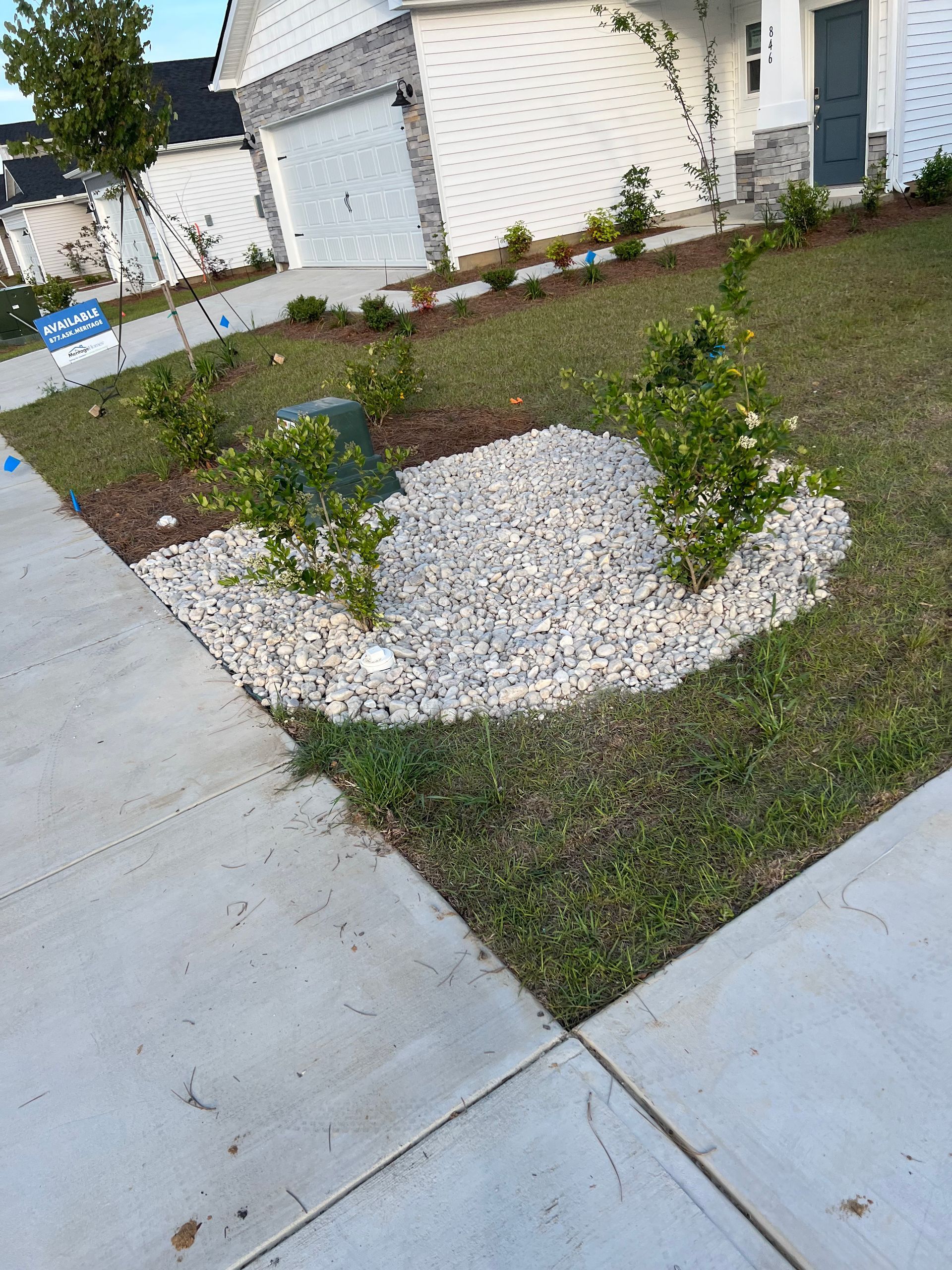 Front yard landscaping with white rock bed, small shrubs, green lawn, and sidewalk. A utility box sits within the rock bed.