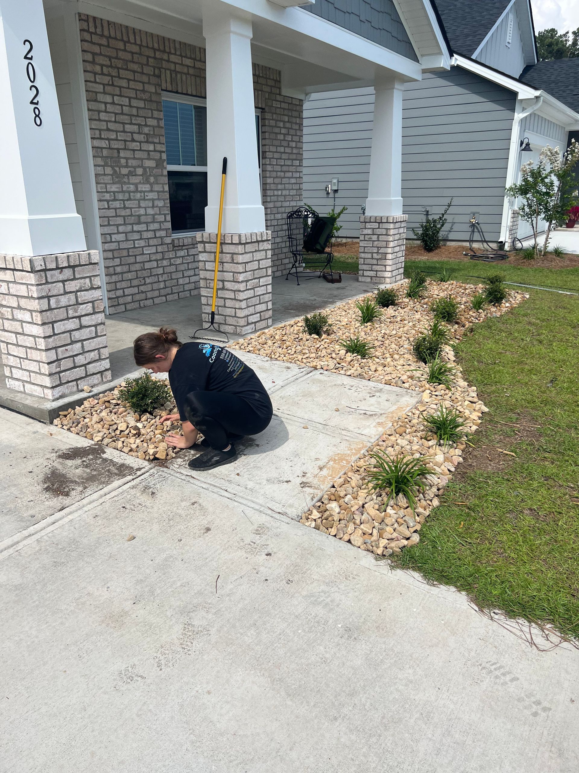 A person tending to plants in a landscaped area next to a concrete walkway in front of a house with stone detailing.